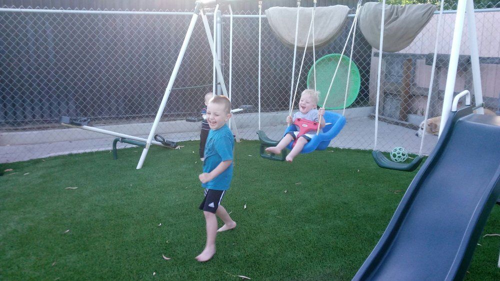 Two children playing on a swing set in a grassy backyard. One child swings, the other laughs.