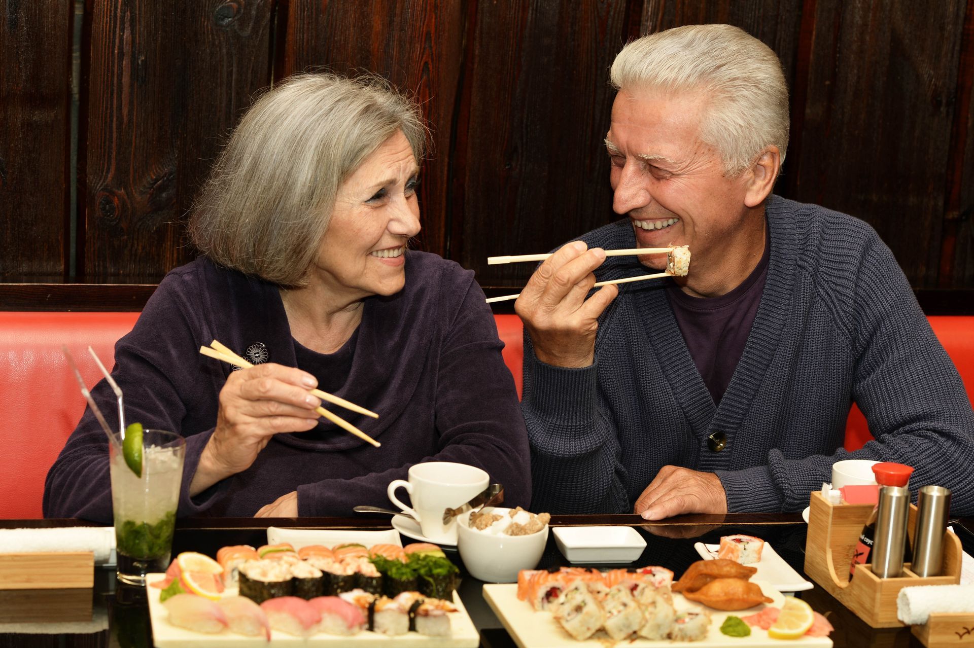 Couple smiling, eating sushi with chopsticks in a restaurant. Red booth, food on the table.