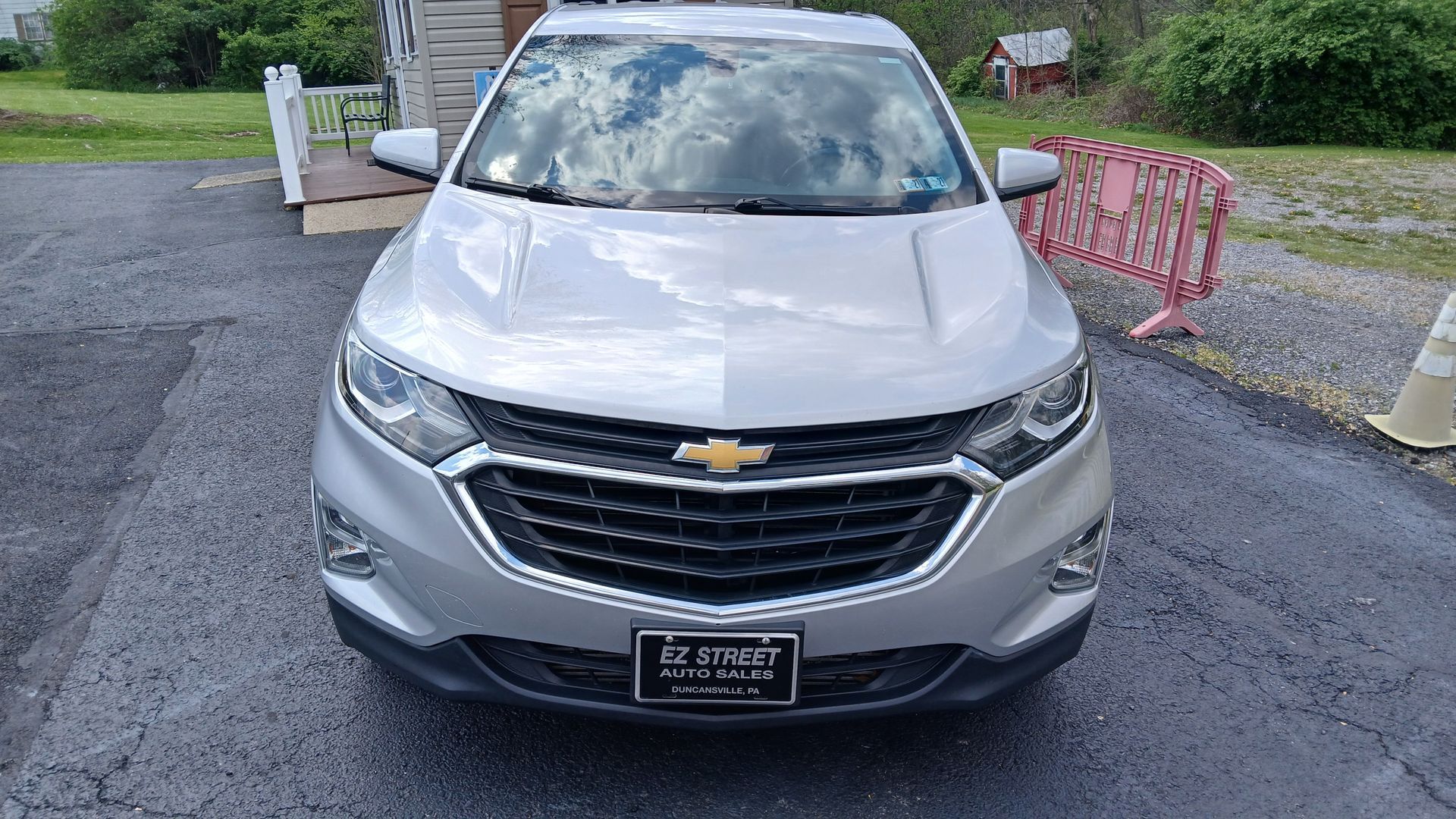 Silver Chevrolet SUV parked on a gravel driveway, front view with cloudy sky reflection on windshield.