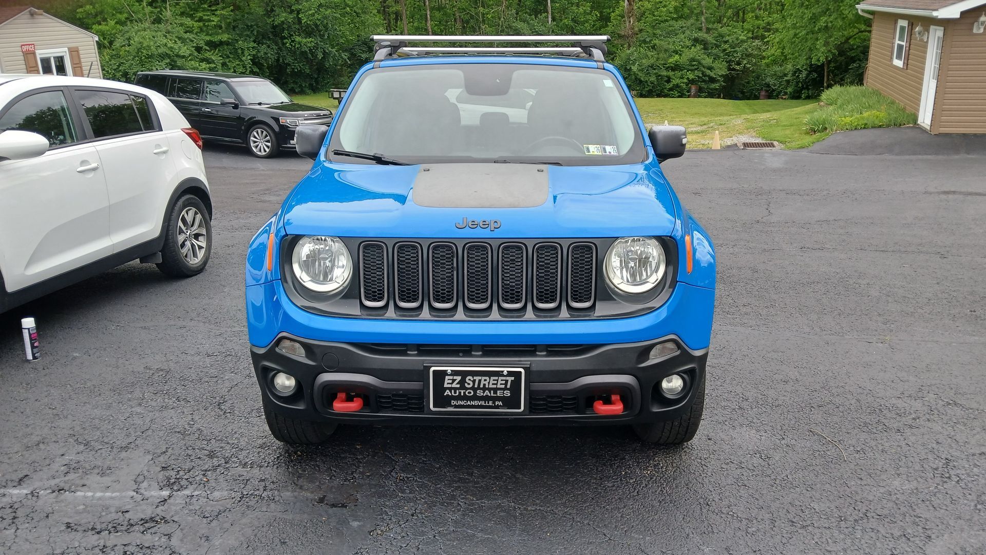 Blue Jeep parked on wet pavement, front view, with other cars and greenery in the background.