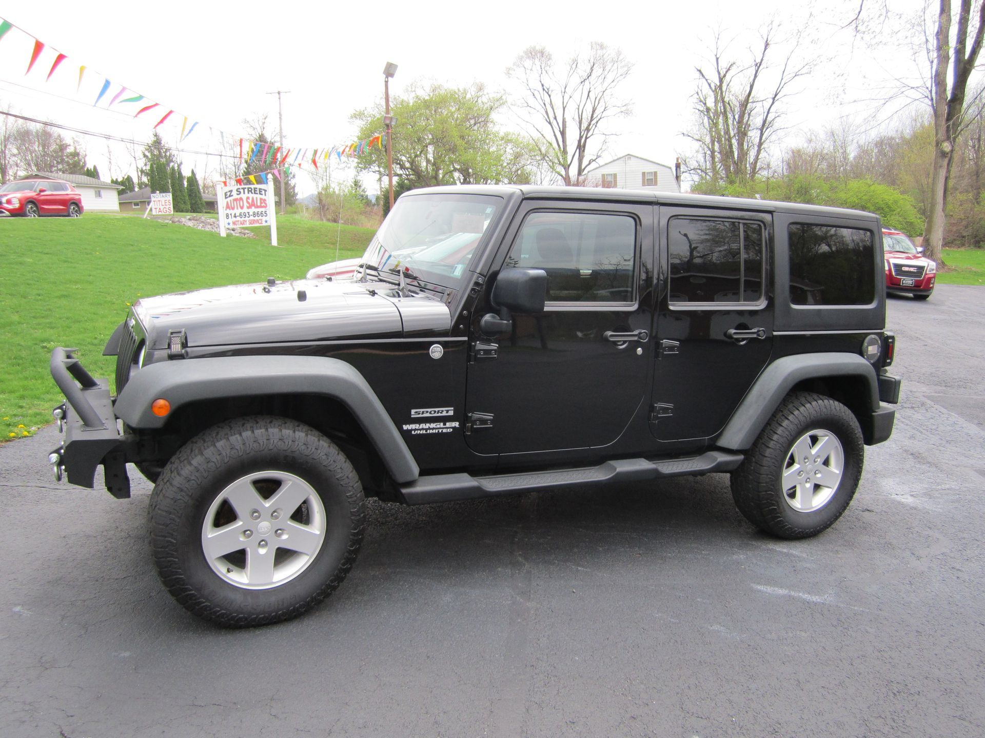 A black Jeep Wrangler Unlimited four-door SUV parked on an asphalt lot with a grassy hill and flags in the background.