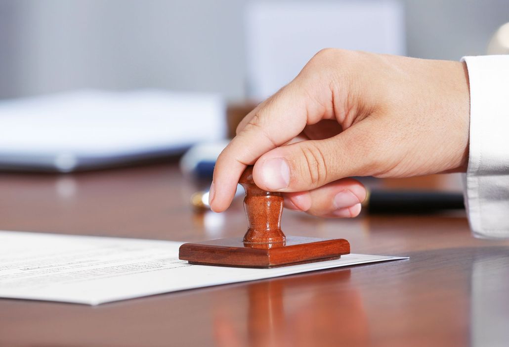 A hand presses a wooden rubber stamp onto a document on a wooden desk.