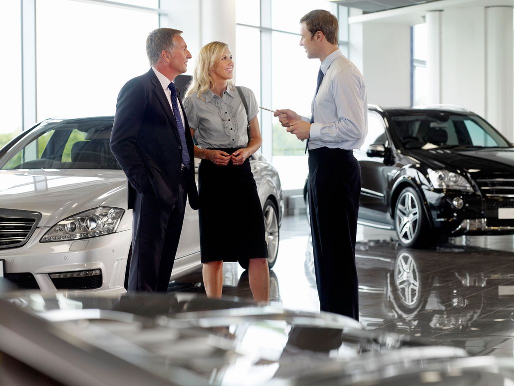 A salesperson speaks with a couple inside a car dealership showroom featuring two luxury vehicles.