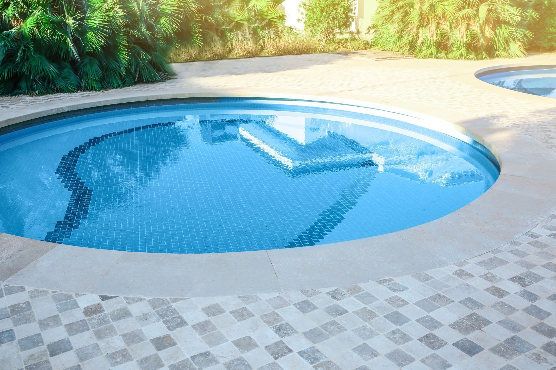 Blue, round pool with steps; surrounded by beige, tiled deck; greenery in background.