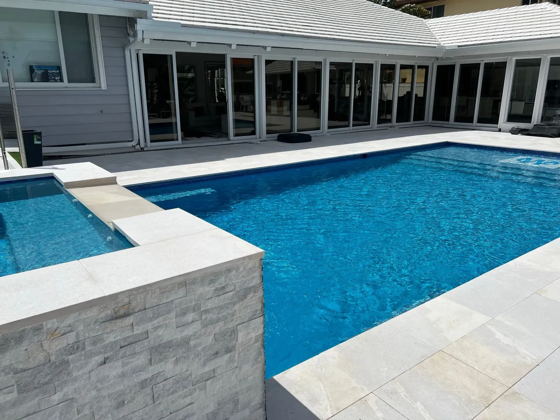 A rectangular swimming pool and spa with blue water surrounded by white stone and a house with sliding glass doors.