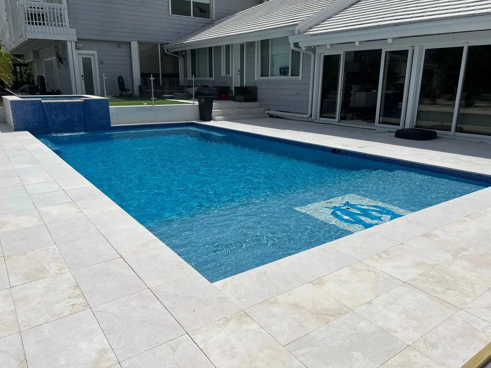 Rectangular pool with blue water and surrounding white tile patio. A house is in the background.