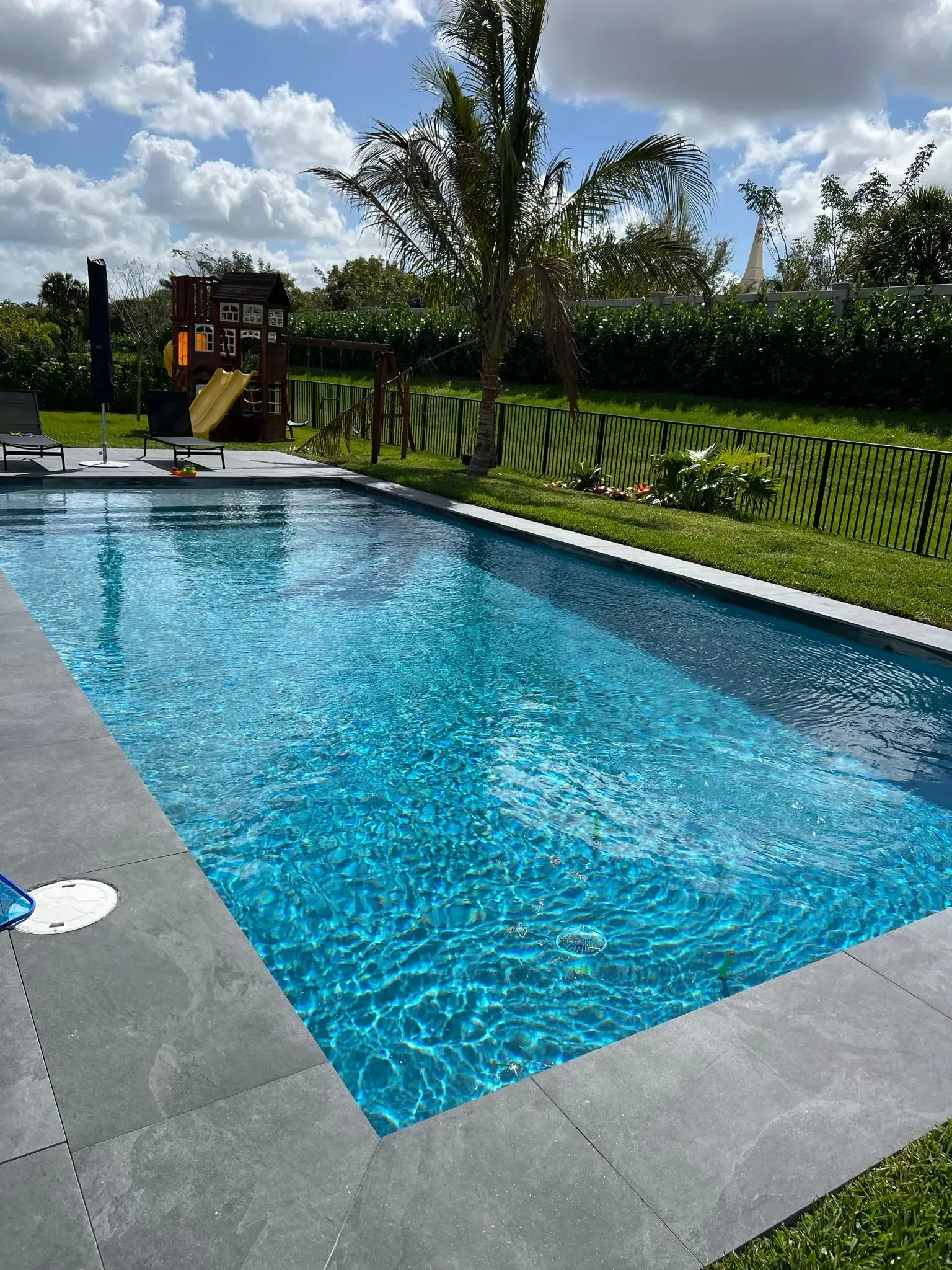 Pool with blue water and gray stone surround; green lawn, trees, and blue sky in background.