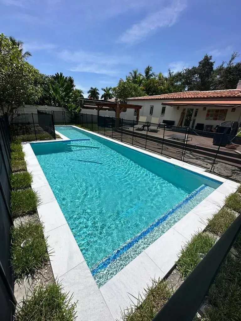 Rectangular swimming pool with clear blue water, surrounded by white tiles and greenery under a sunny sky.