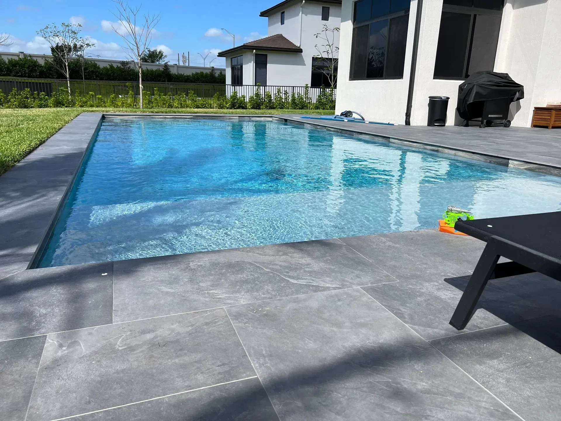 Rectangular pool with blue water and gray stone patio next to a modern white house under a sunny sky.