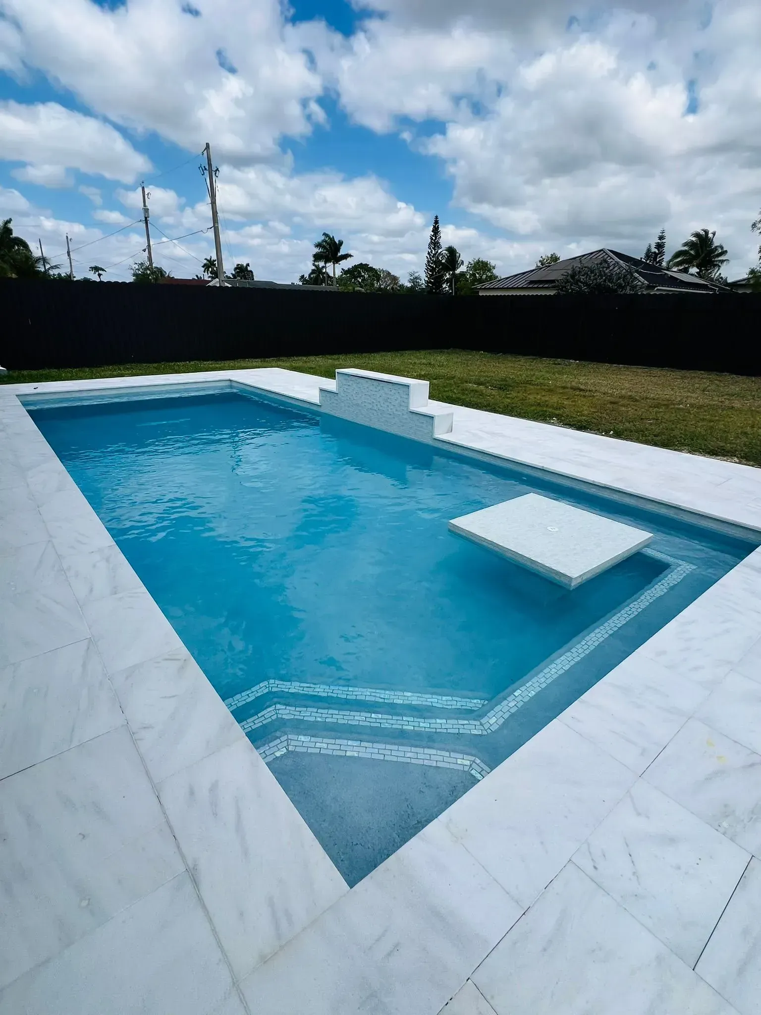 A rectangular swimming pool with white tile surround. Blue water, steps, and a white sunbathing platform.