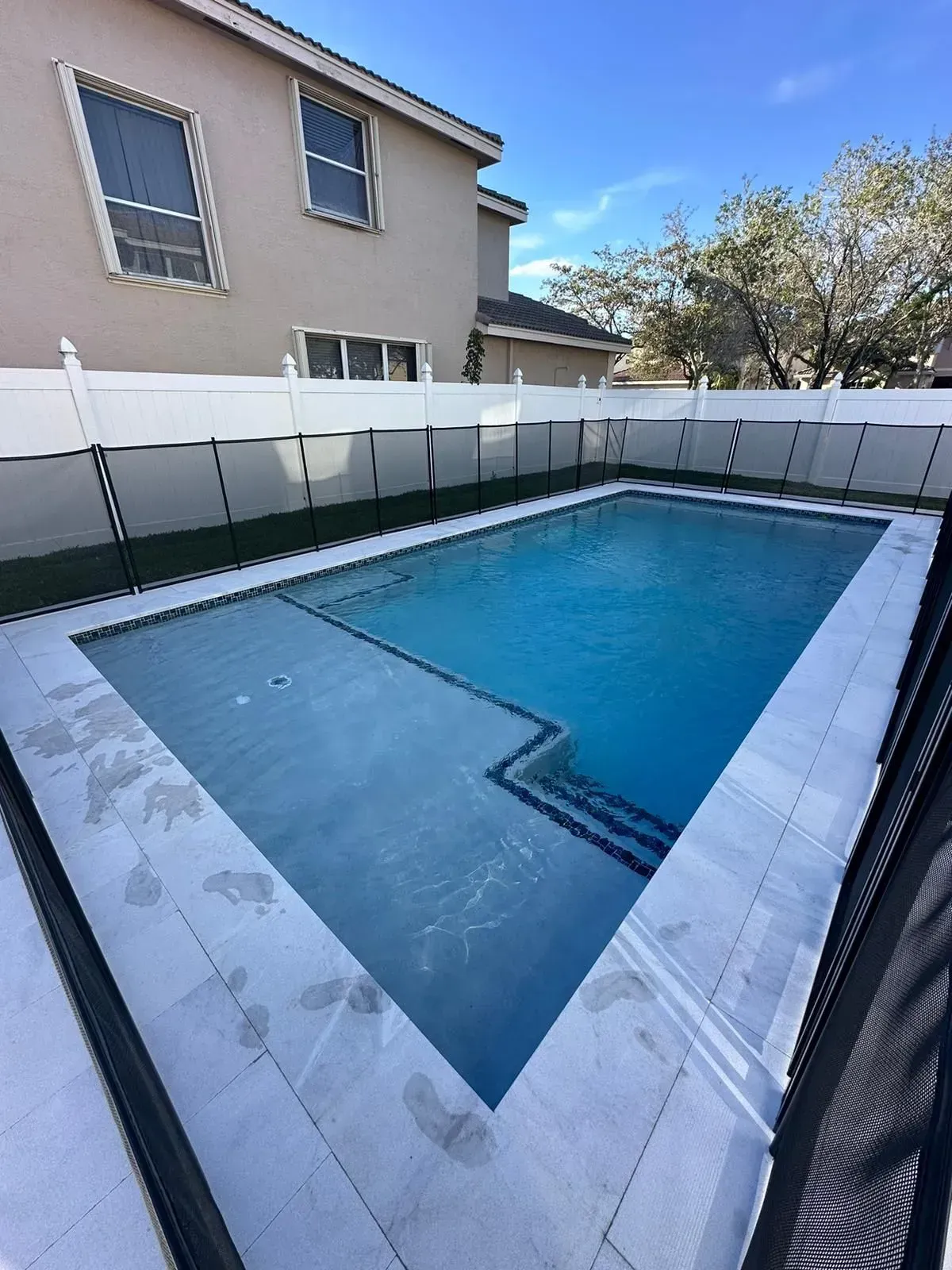 A rectangular swimming pool surrounded by a white fence and a two-story beige house on a sunny day.