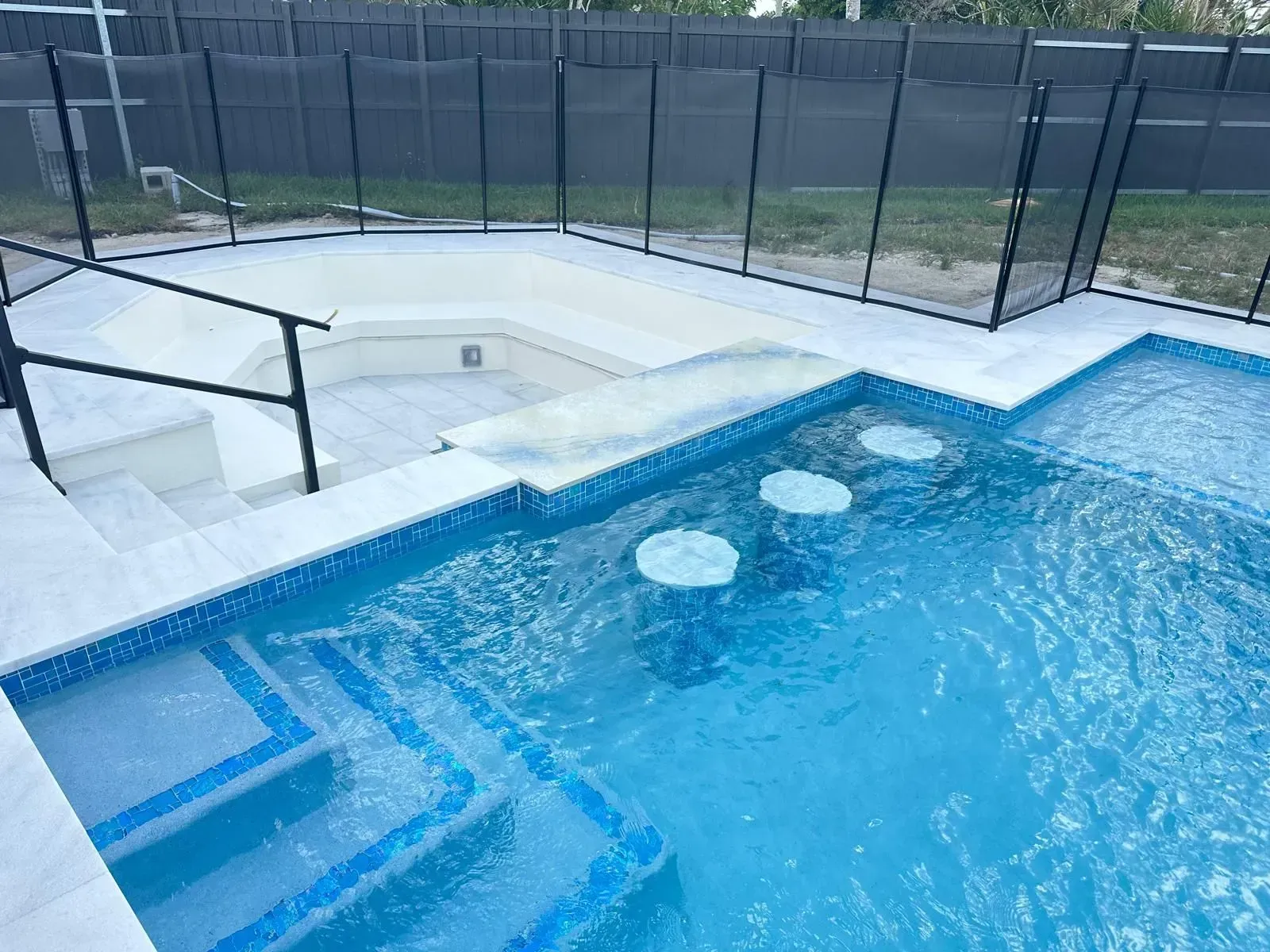 Swimming pool with built-in bar stools and steps. Blue water, white tiling, black safety fence.