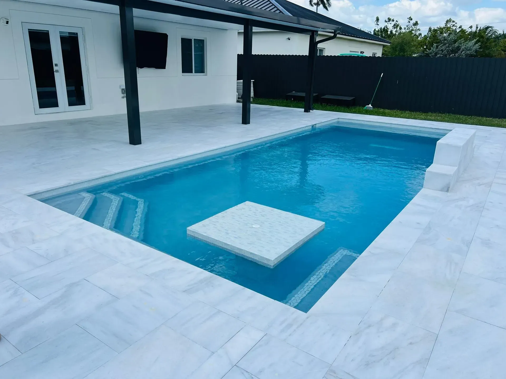 Swimming pool with white marble-like tiles and a floating square table, adjacent to a house.
