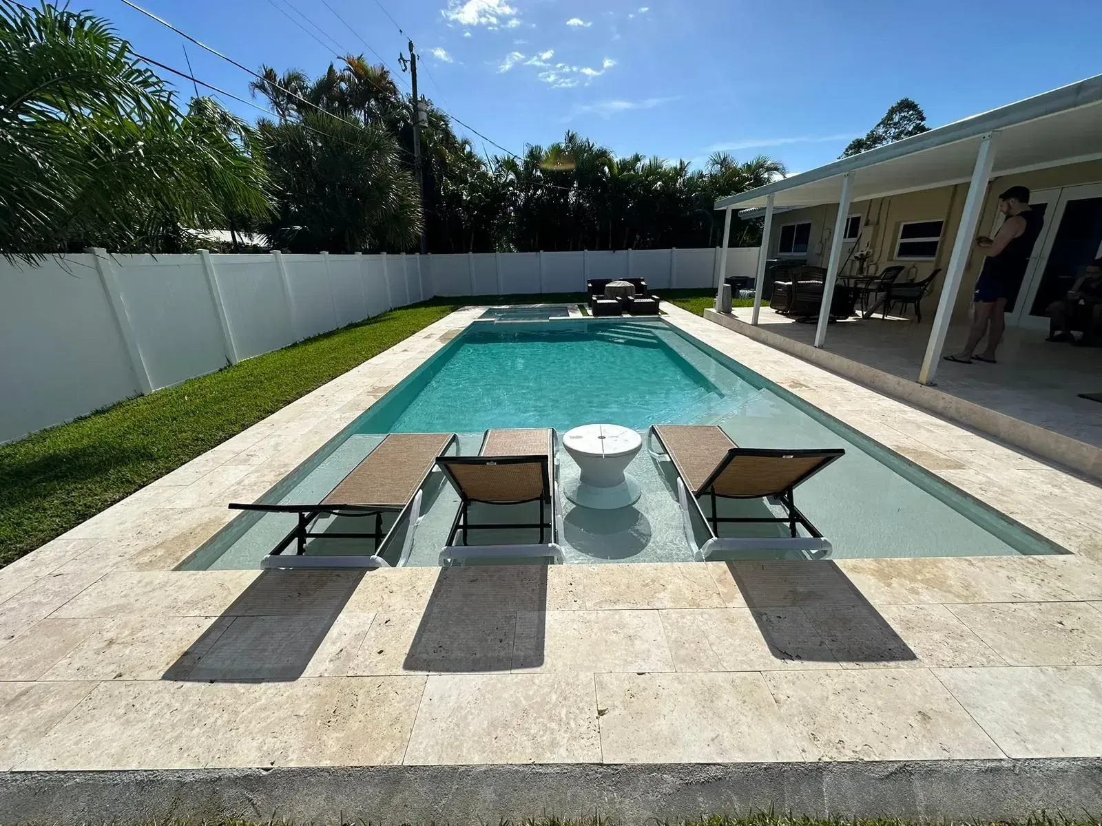Swimming pool with chaise lounges in shallow water; white fence and covered patio in background.