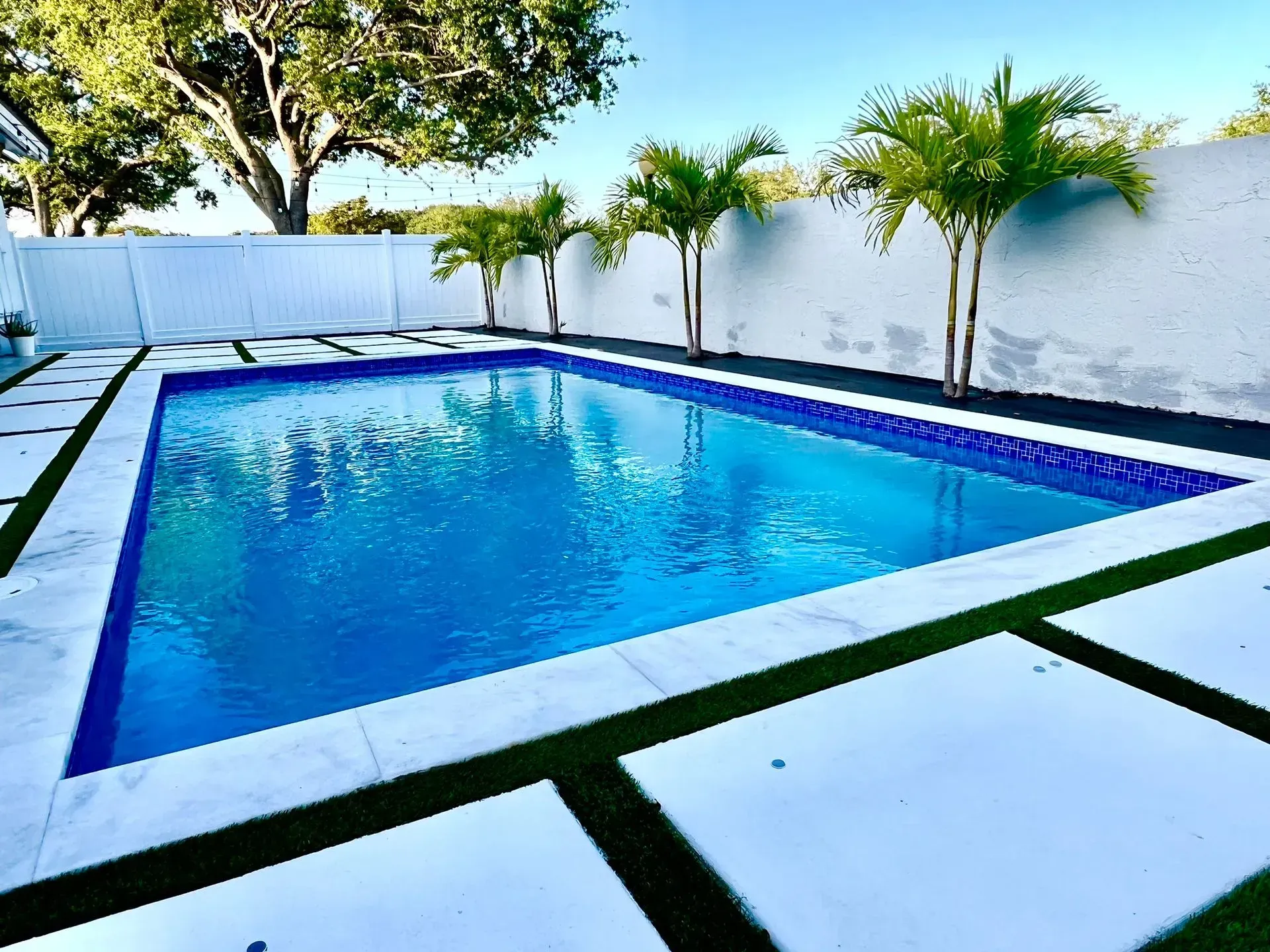 Rectangular pool with blue water and marble edge, bordered by artificial grass and white walls with palm trees.