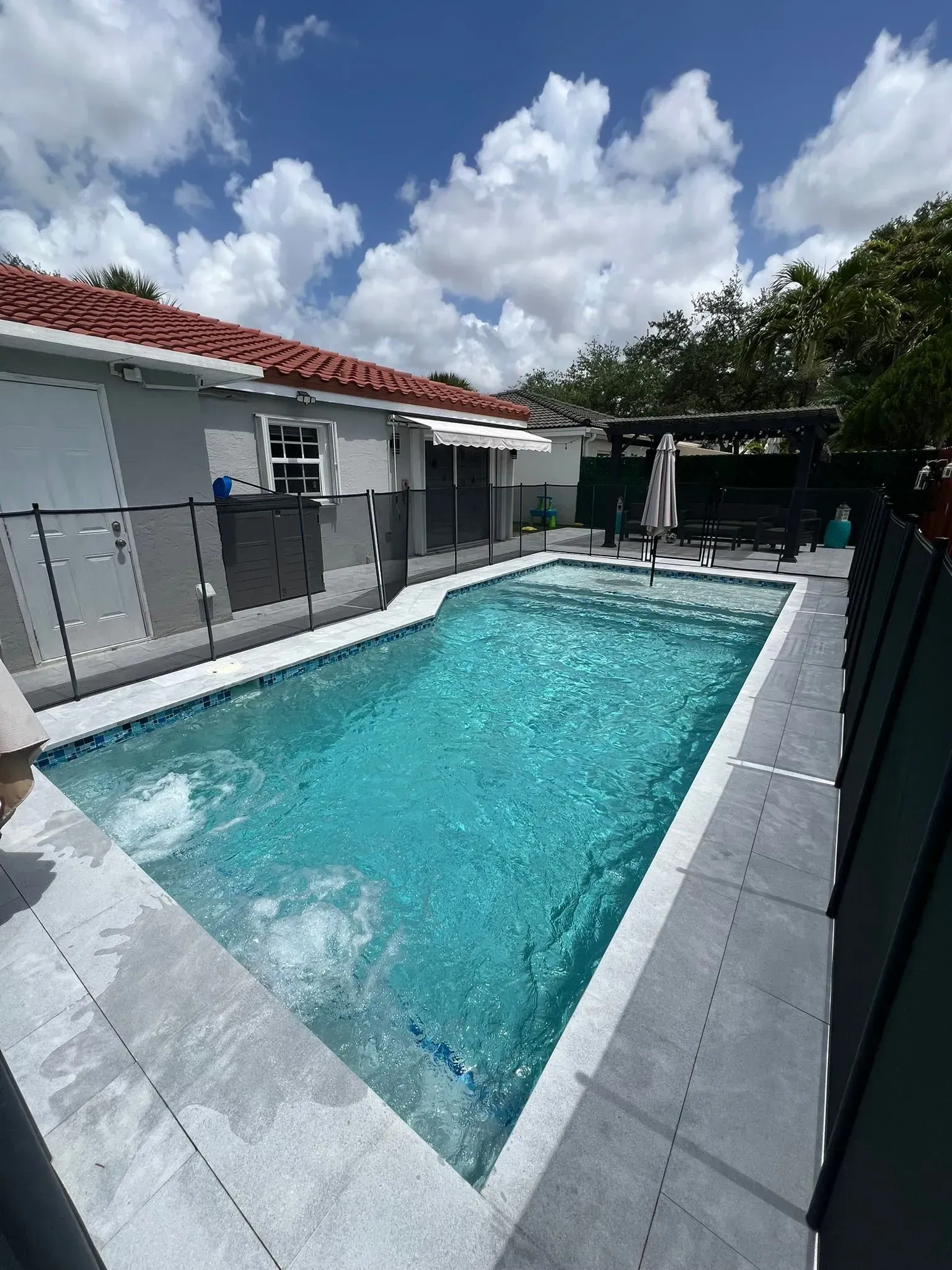Swimming pool with blue water surrounded by a gray patio, fence, and a building under a cloudy sky.