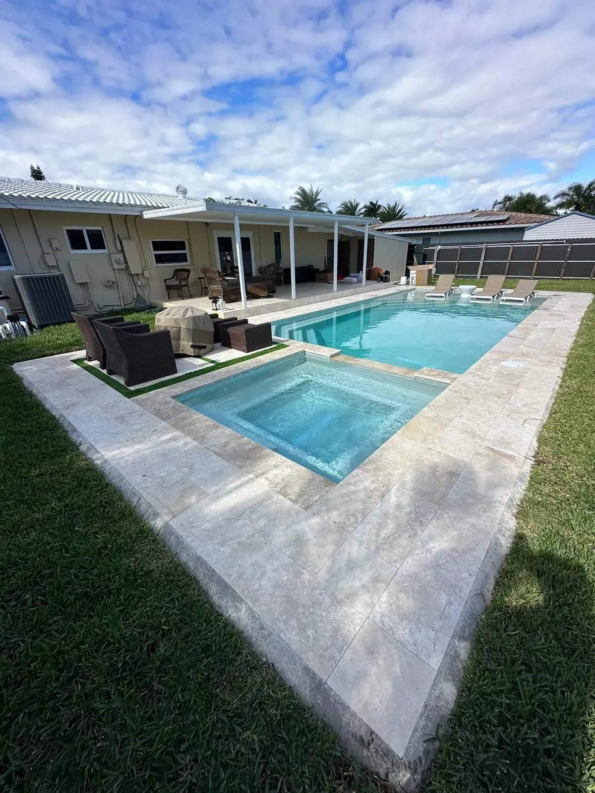 Swimming pool and spa surrounded by light-colored stone, with a covered patio in the background.