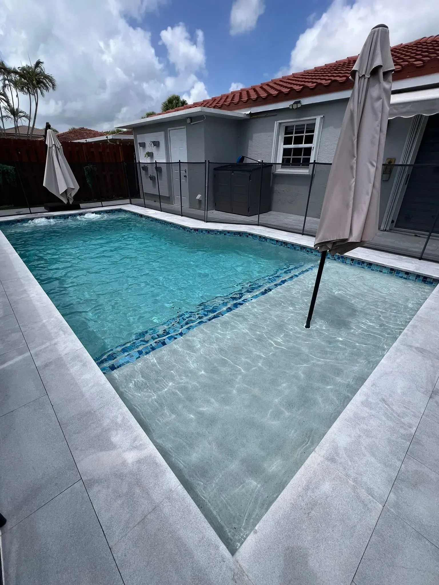 Swimming pool with light blue water and steps, umbrellas, and a house in the background.