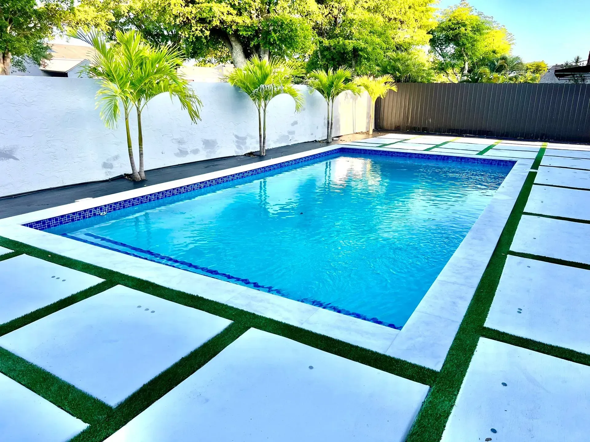Rectangular swimming pool with blue water surrounded by white pavers and green turf.
