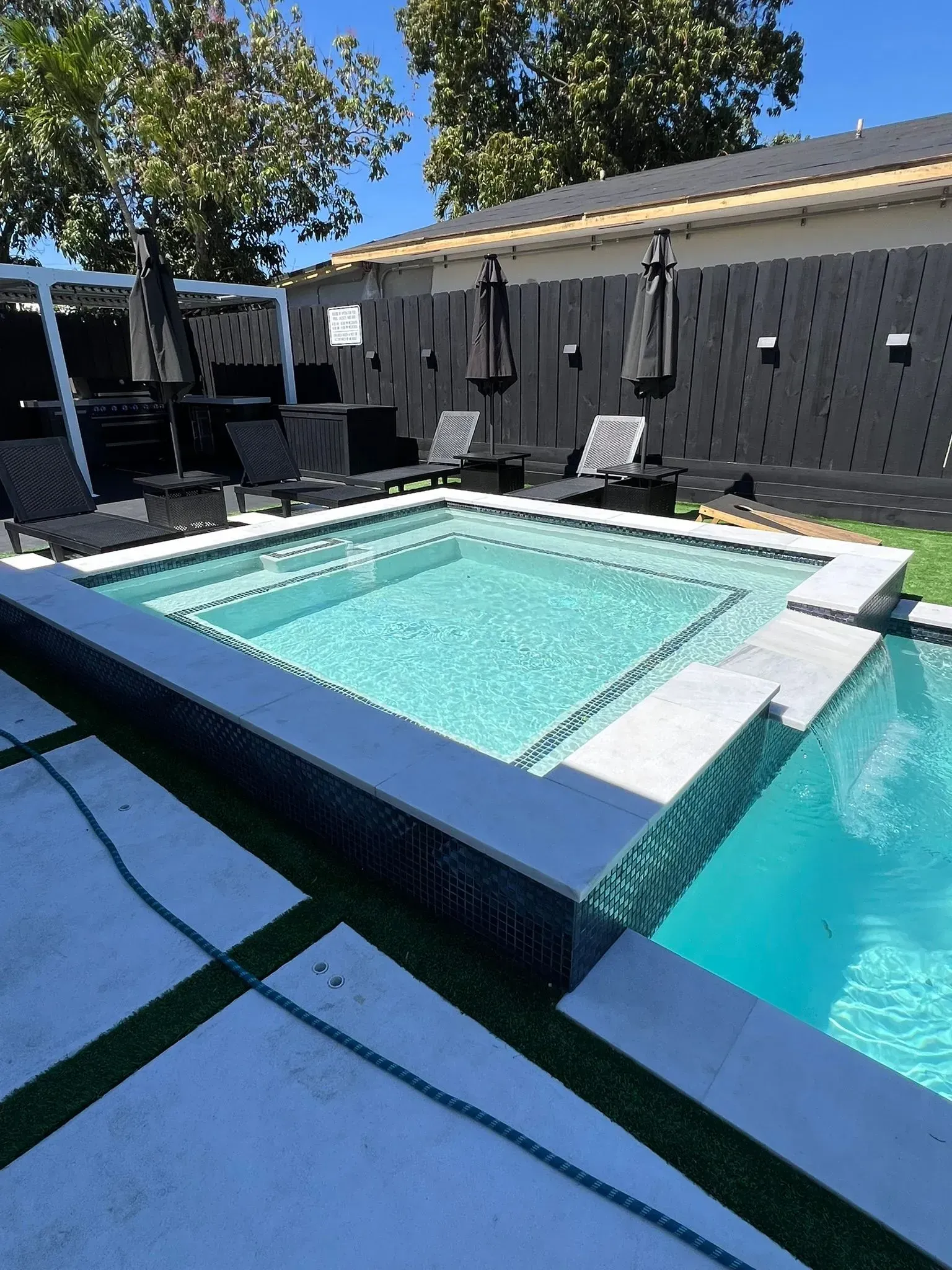 Swimming pool with attached jacuzzi. Black and white lounge chairs and umbrellas. Black fence.