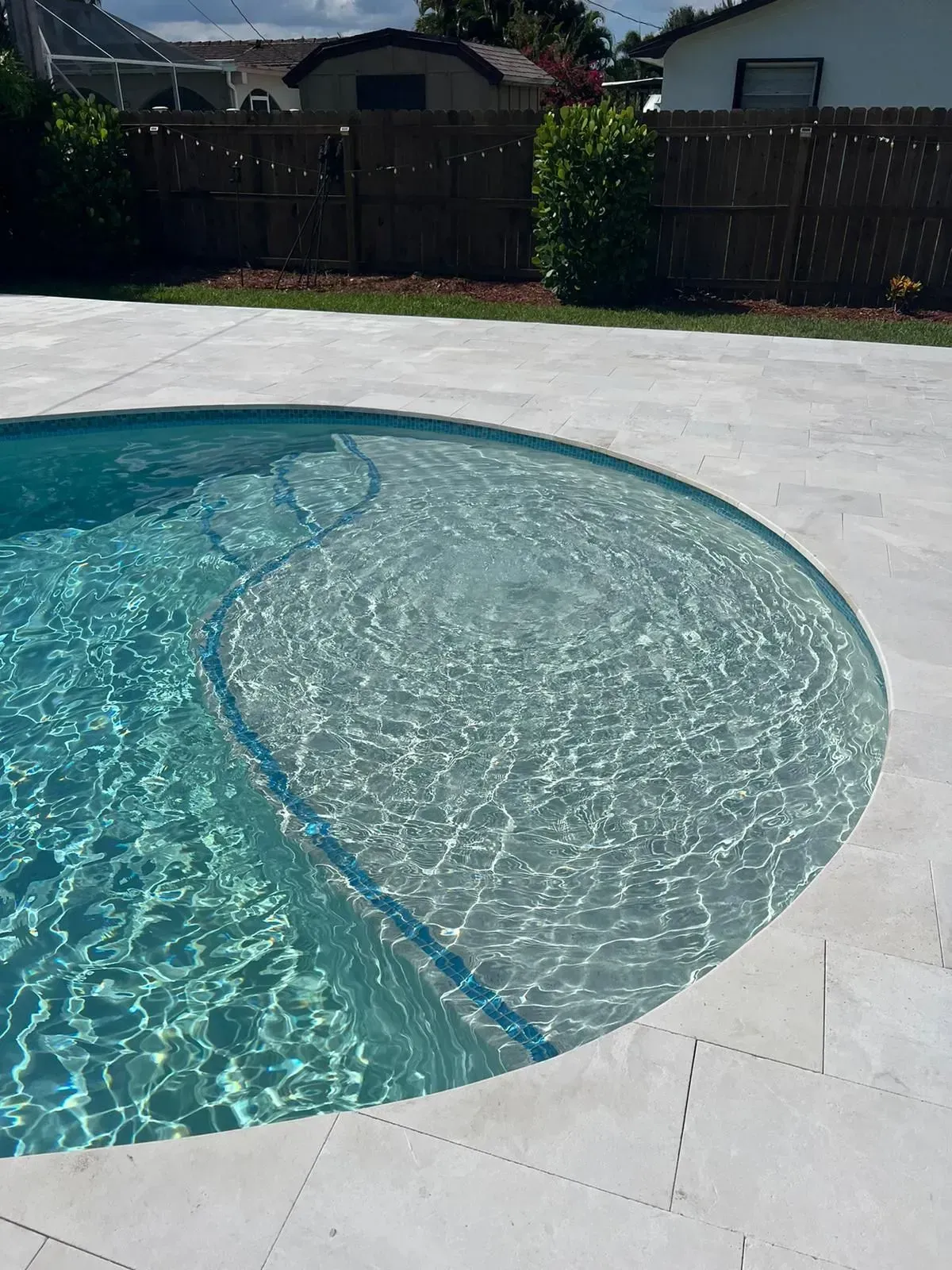 A pool with blue-green water and a light-colored stone deck, surrounded by grass and a fence.
