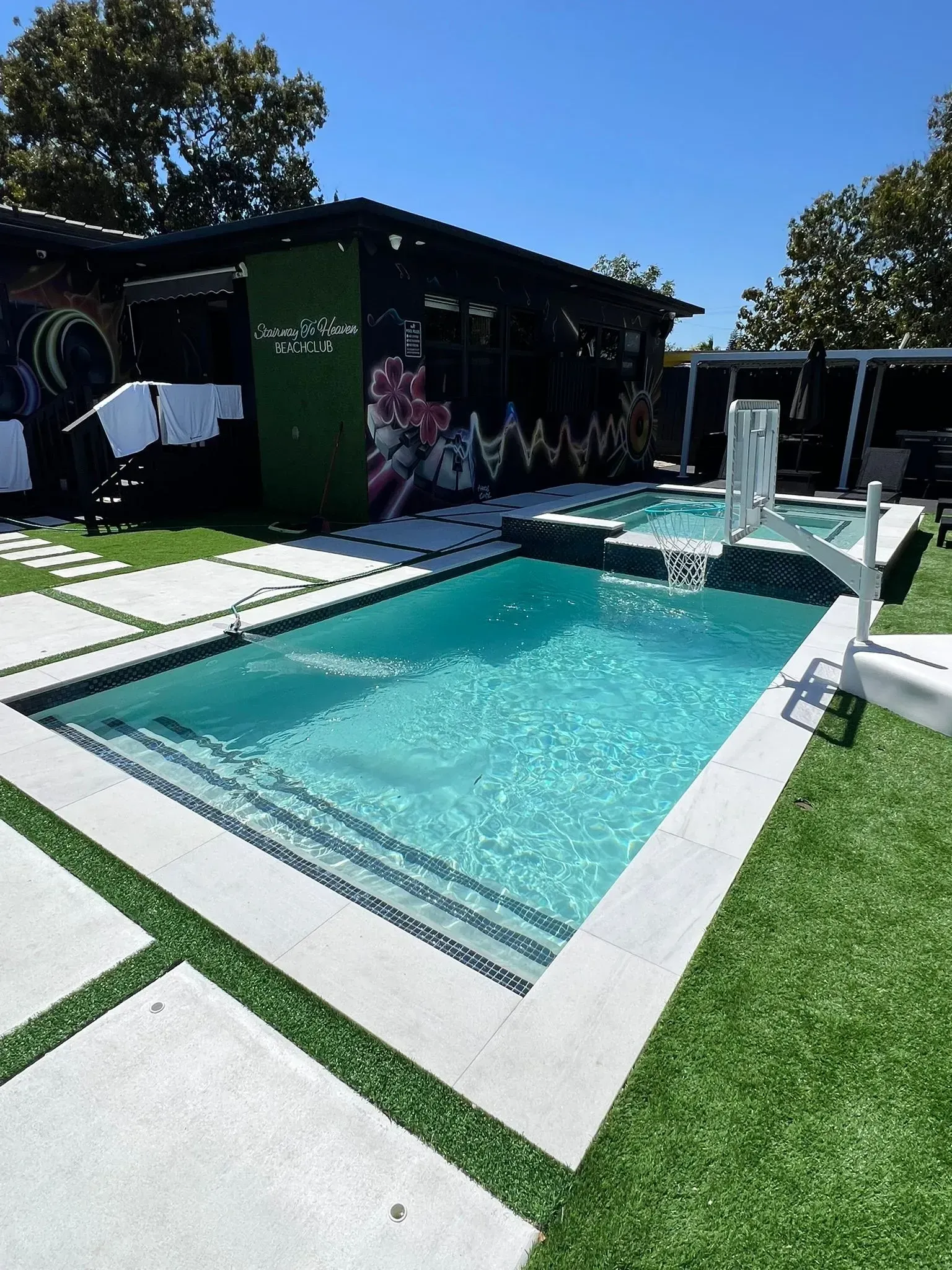 Pool with steps, next to a smaller pool, patio, and building with a mural in a yard.