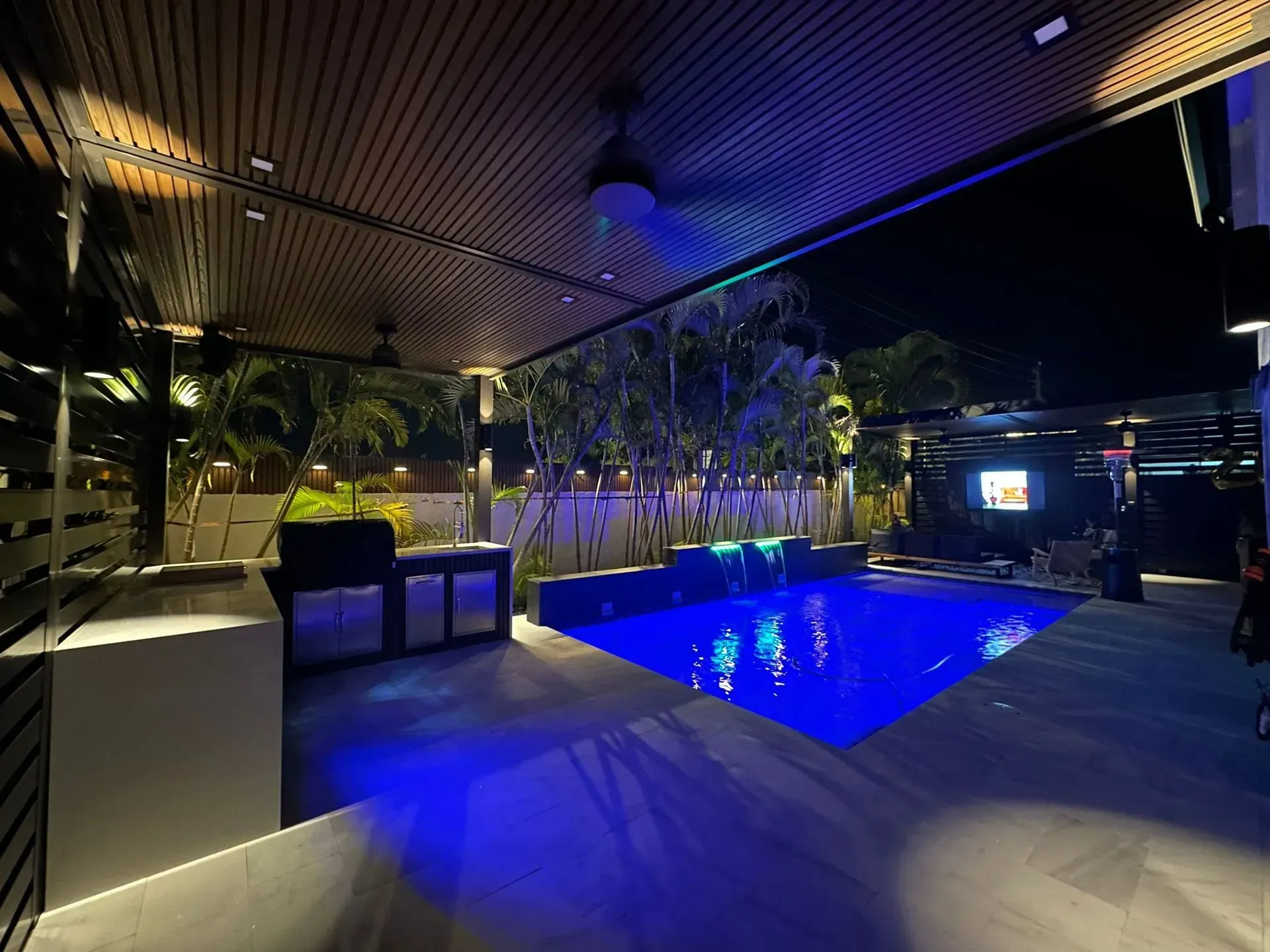 Outdoor night scene: Pool with blue lights, outdoor kitchen, covered patio with dark wood ceiling.