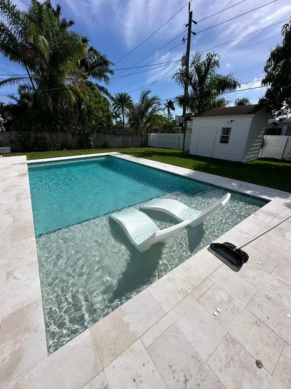 Rectangular pool with white lounge chair, surrounded by light-colored stone and grass, under a blue sky.