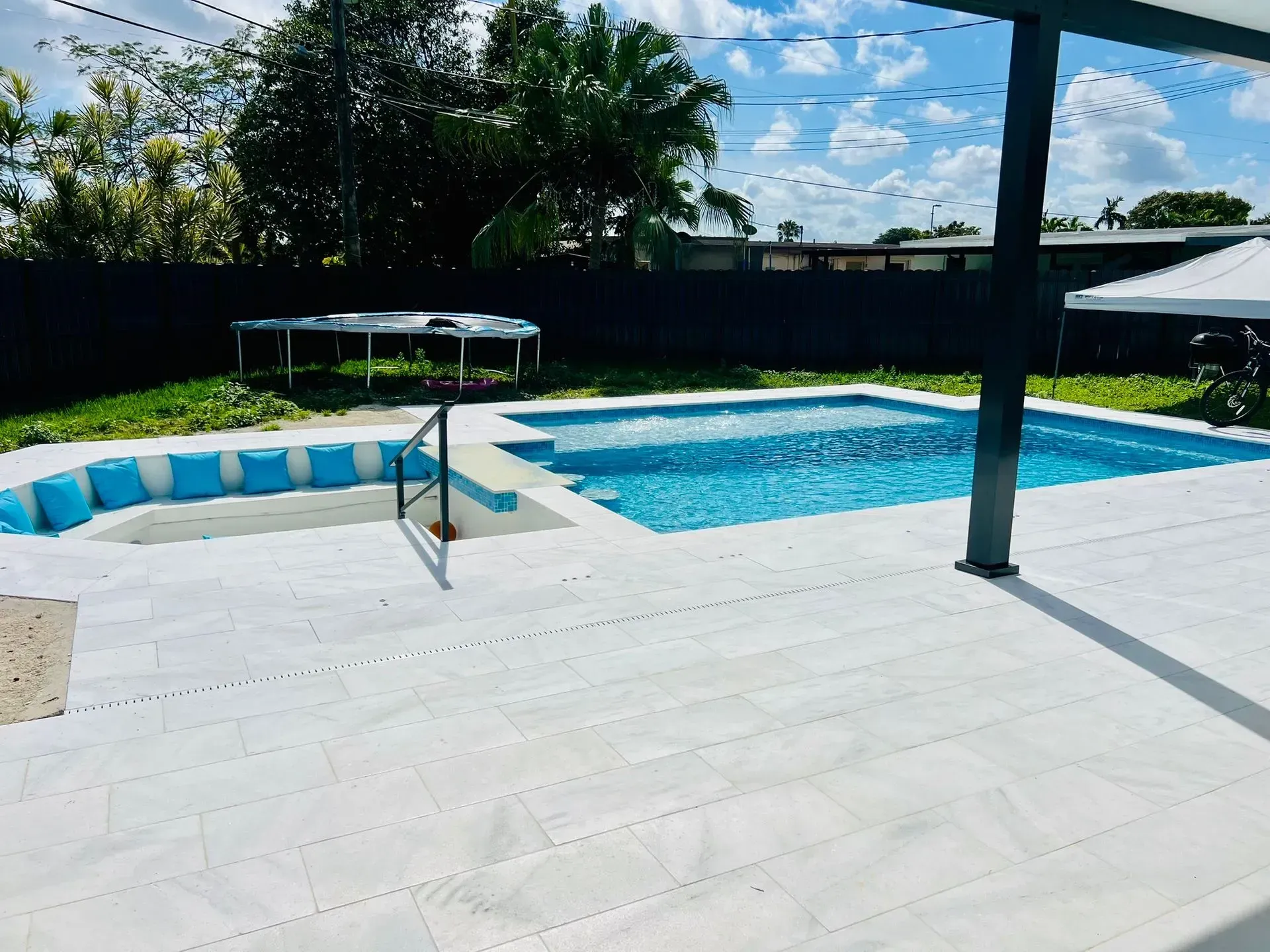 Backyard with pool, white patio, and trampoline. Blue pool water, blue and white cushions. Sunny day.