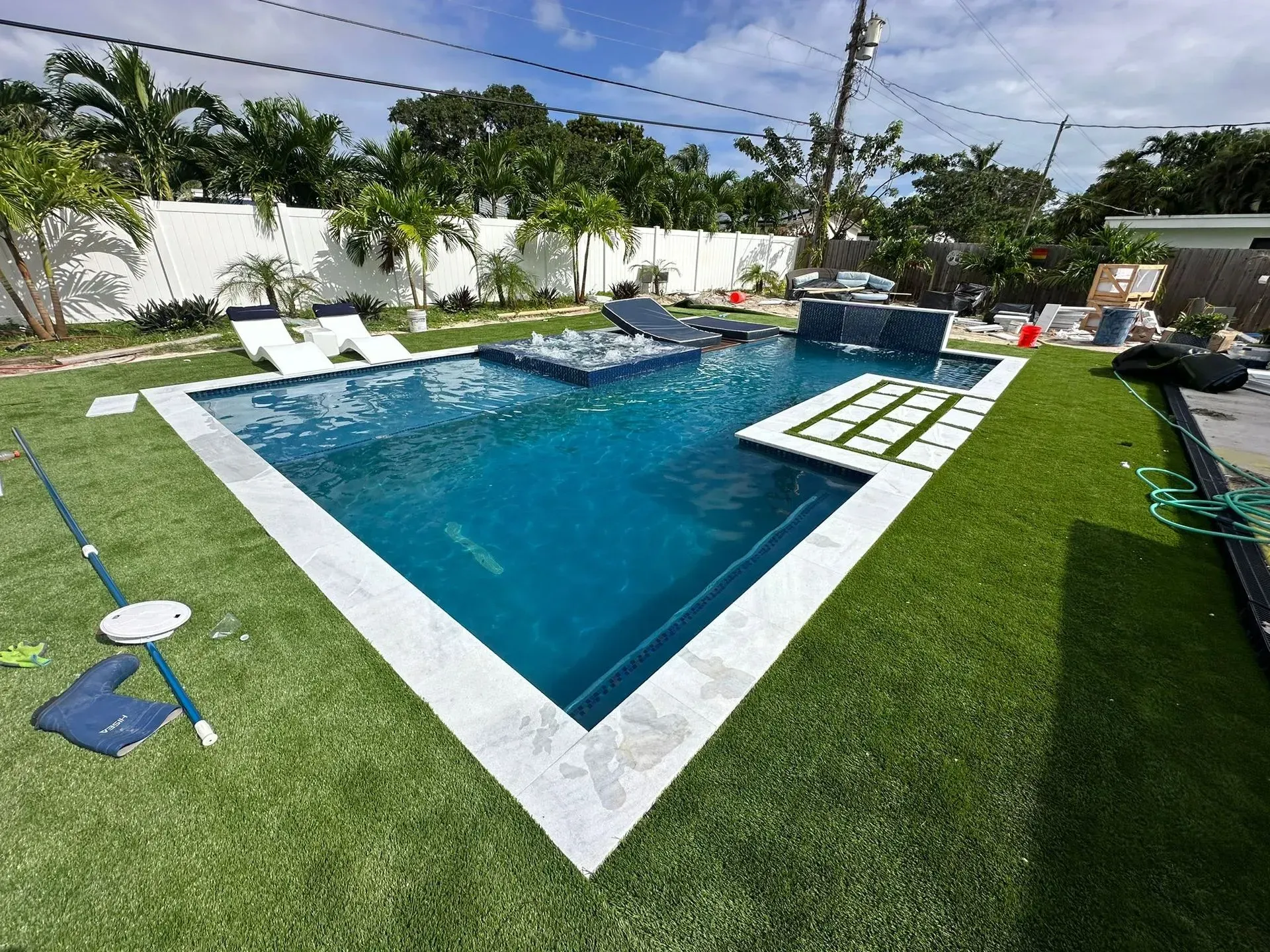 Triangular blue pool with white border, surrounded by green artificial grass. Palm trees and a white fence are in the background.