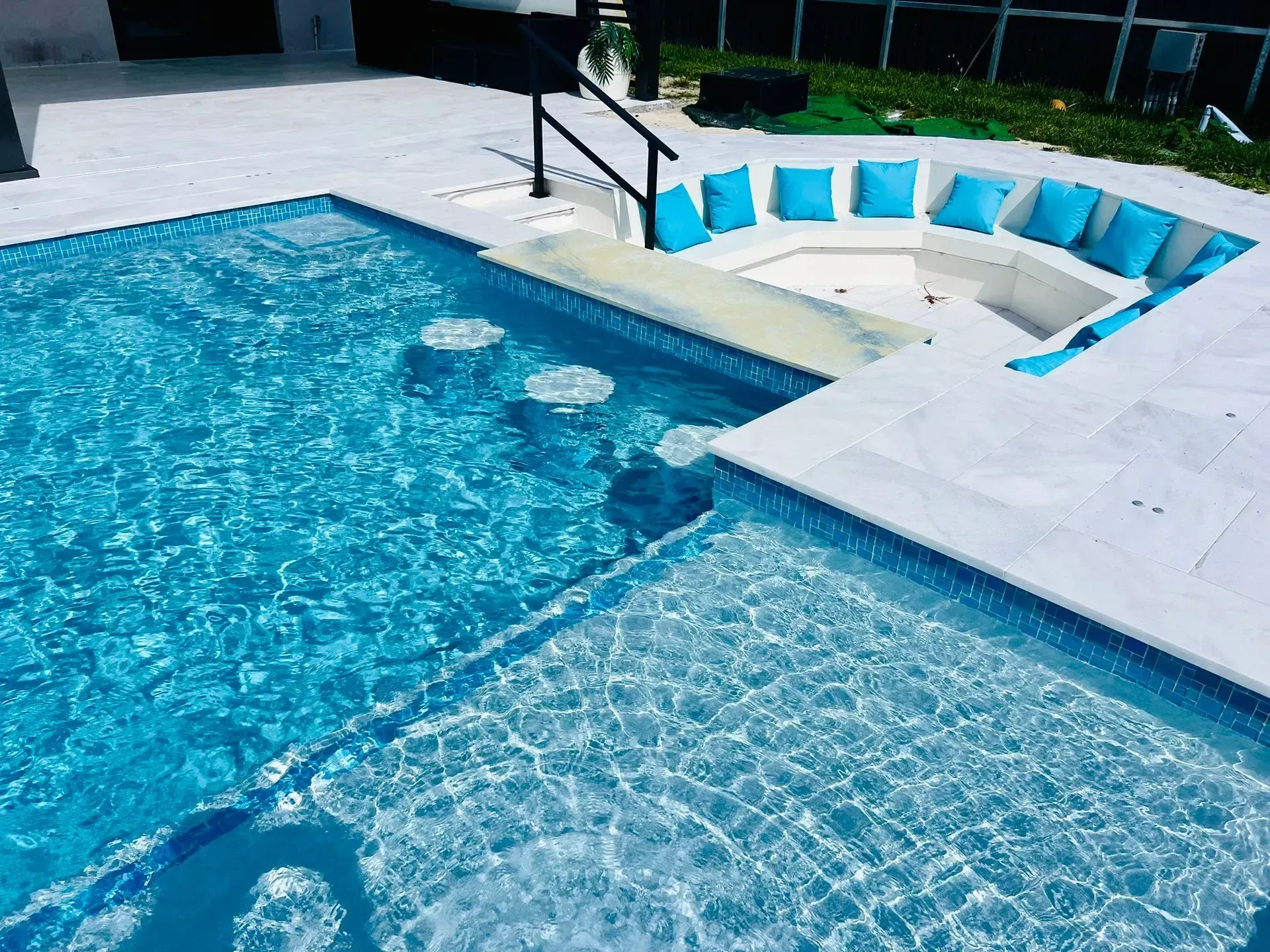 A clear blue swimming pool with built-in seating area and blue cushions. White tile surround.