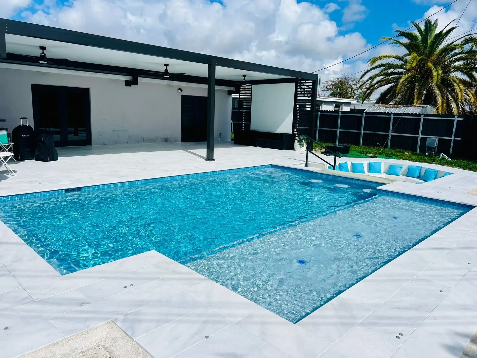 Pool with turquoise water, patio with dark pergola, white tile. Palm tree in background, sunny day.