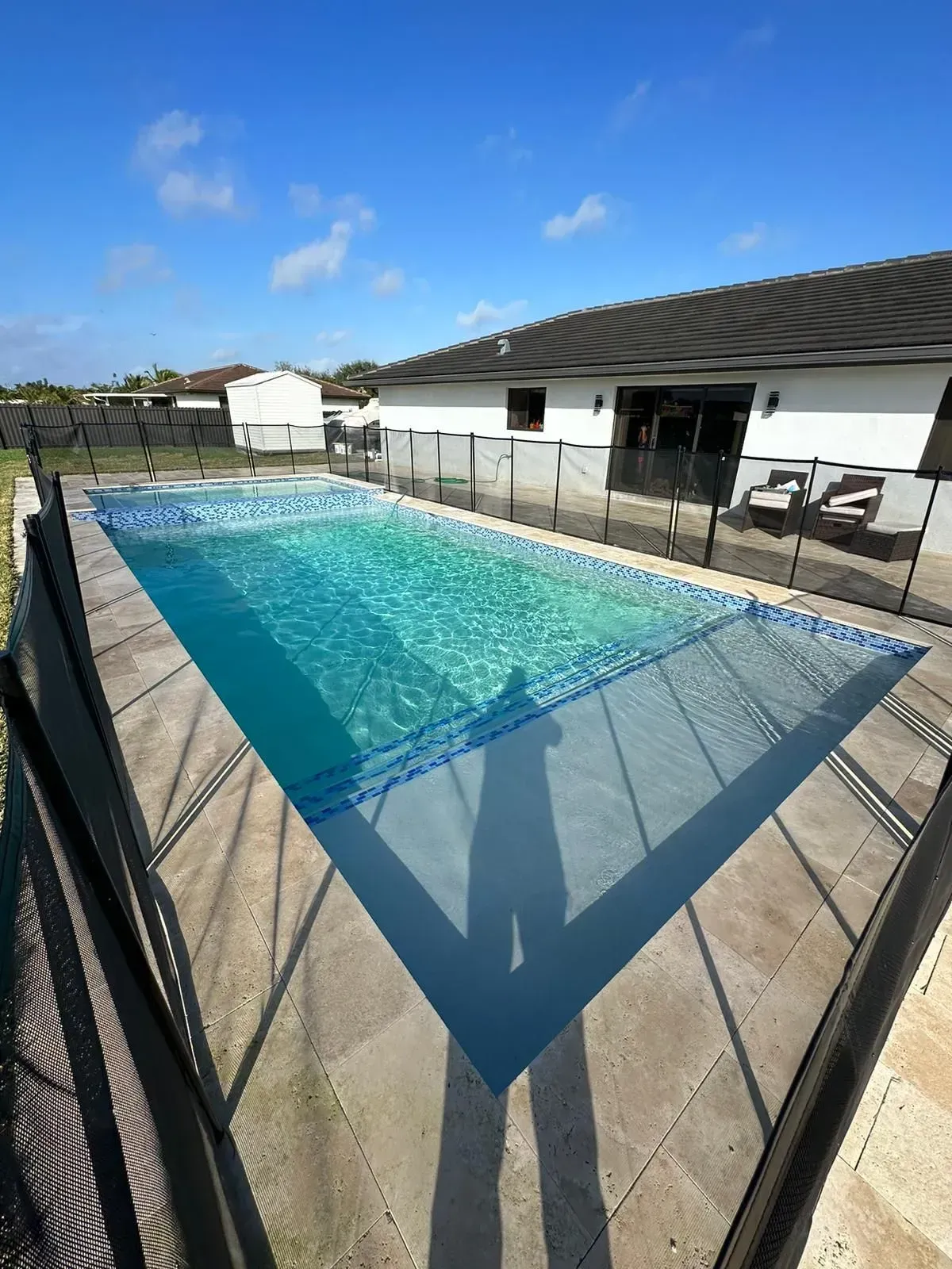 Rectangular swimming pool with clear water, surrounded by a light-colored patio and black fencing, under a blue sky.