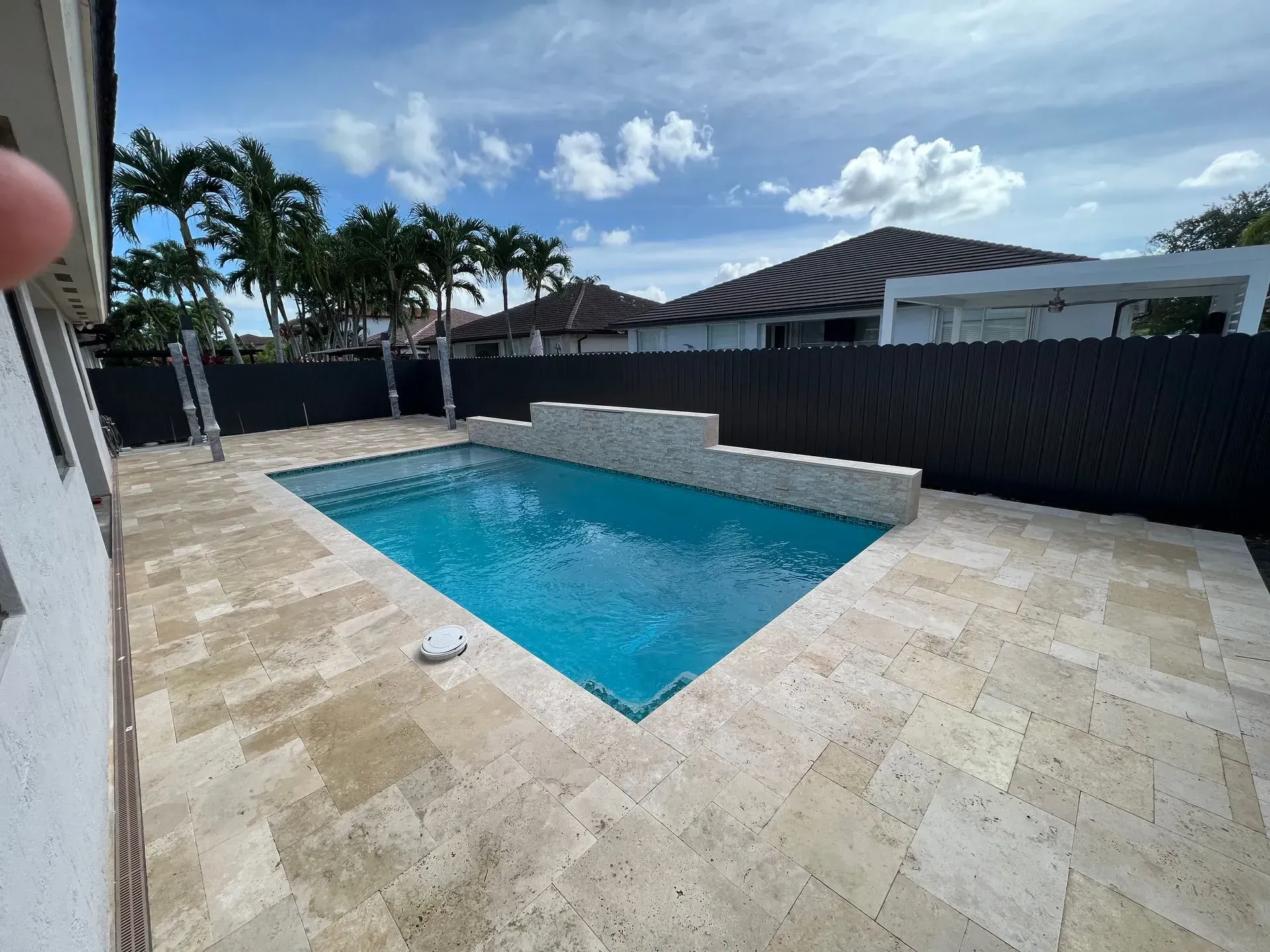A rectangular pool surrounded by light stone tiles. Black fence and palm trees in the background. Bright blue sky.