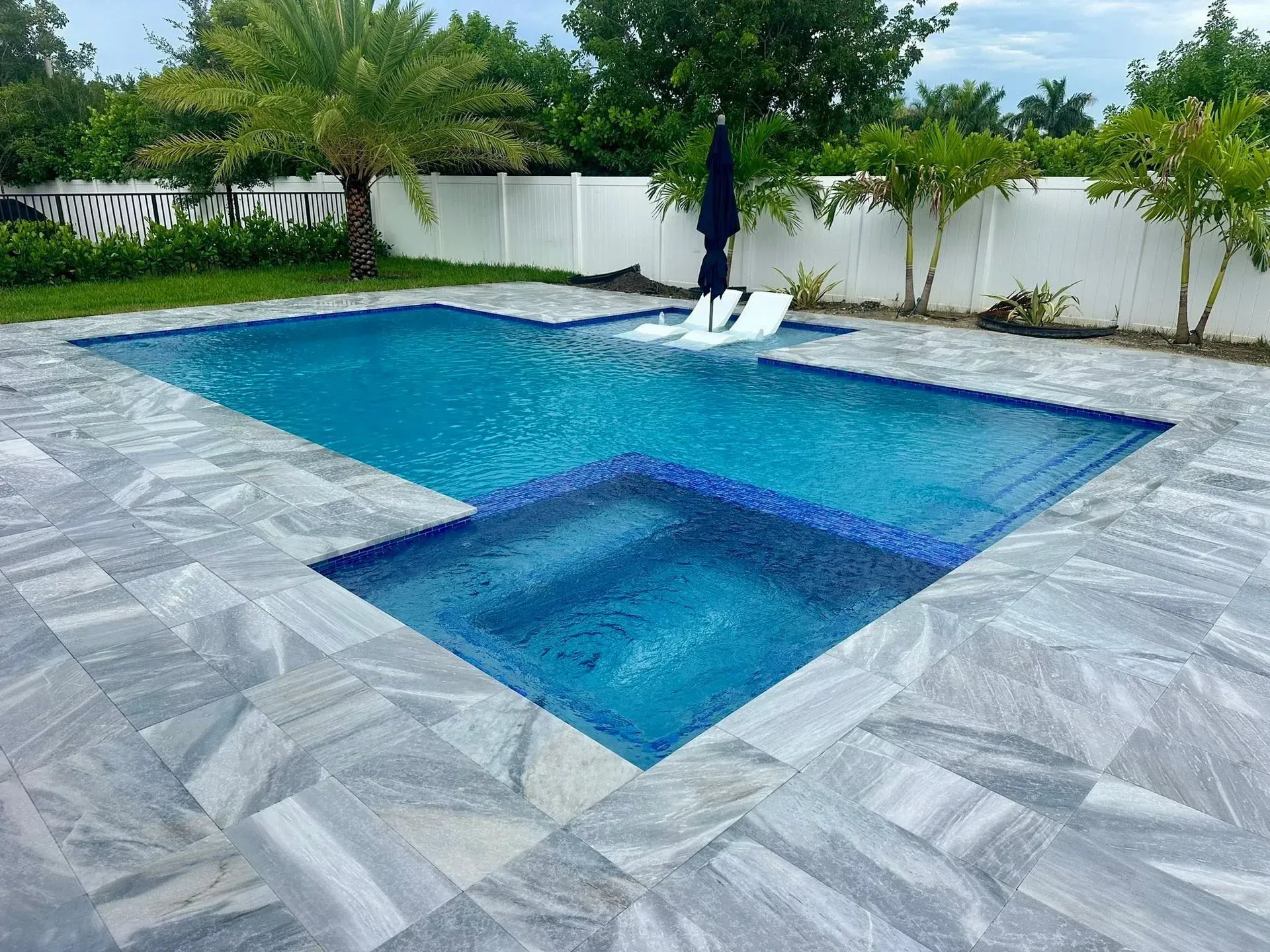 Swimming pool with blue water and gray stone patio surrounded by palm trees and white fence.