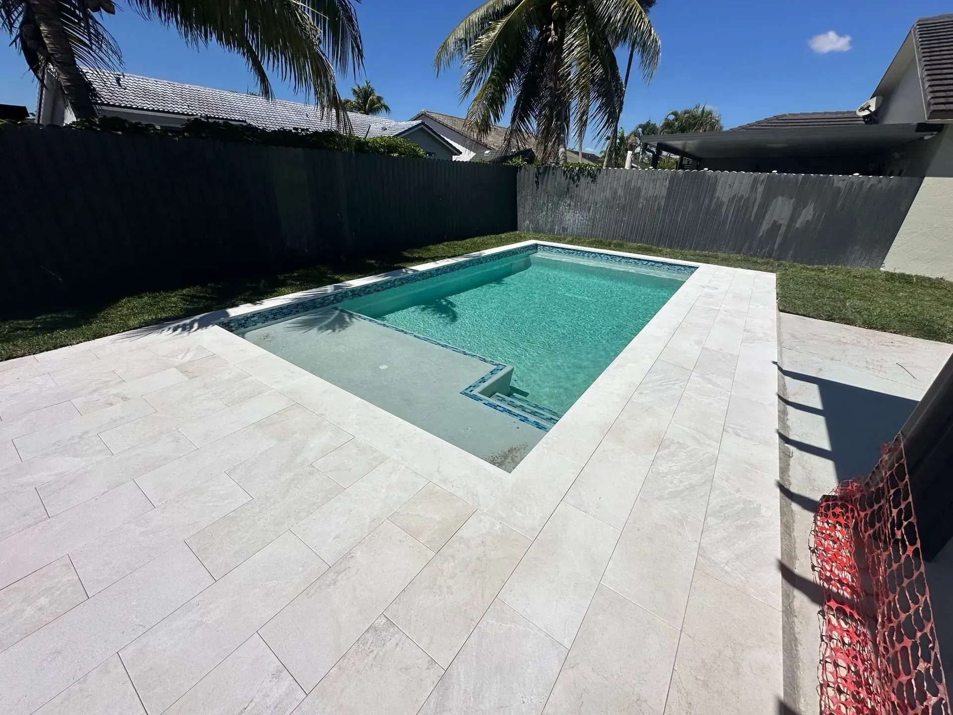 A rectangular swimming pool surrounded by light-colored pavers in a backyard with a dark fence.