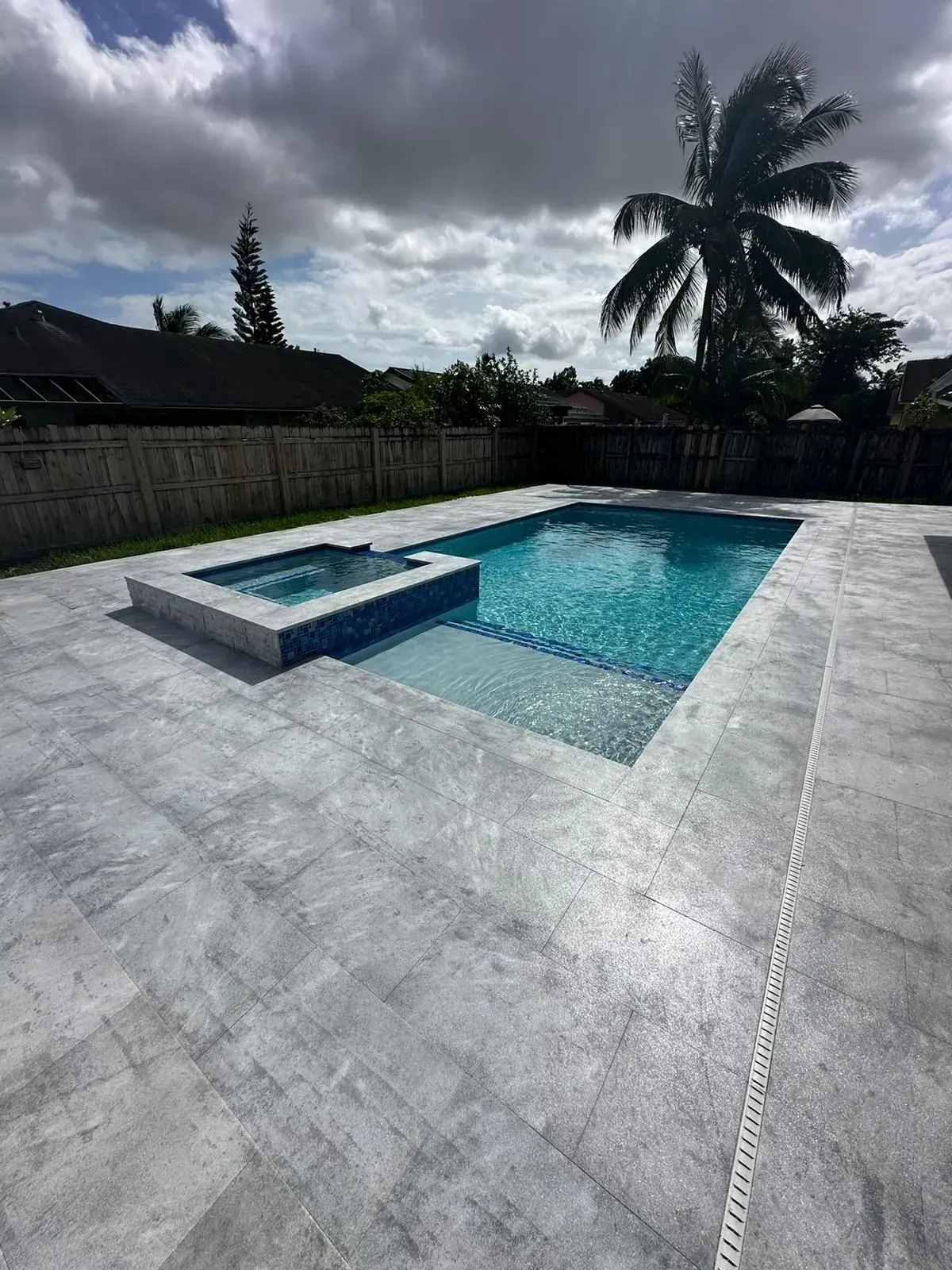 Swimming pool and jacuzzi with gray tiled patio under a cloudy sky.