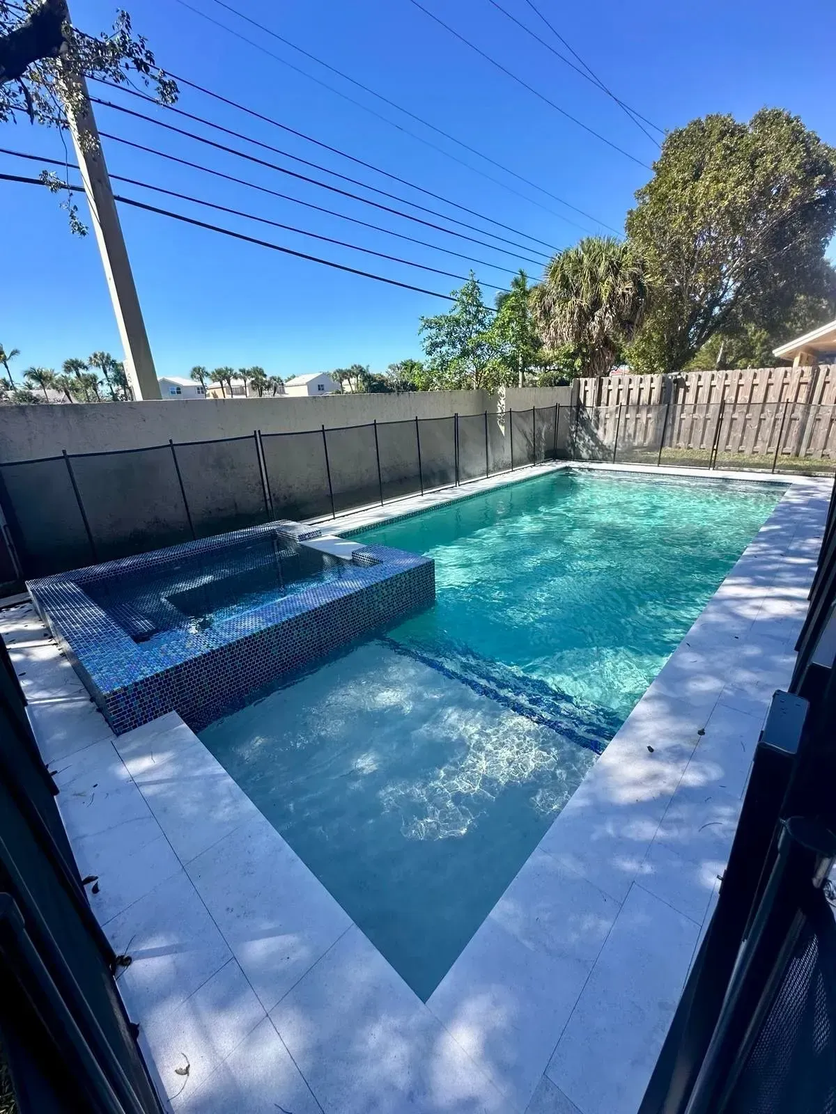 Rectangular pool and attached spa, surrounded by a fence, against a bright blue sky.