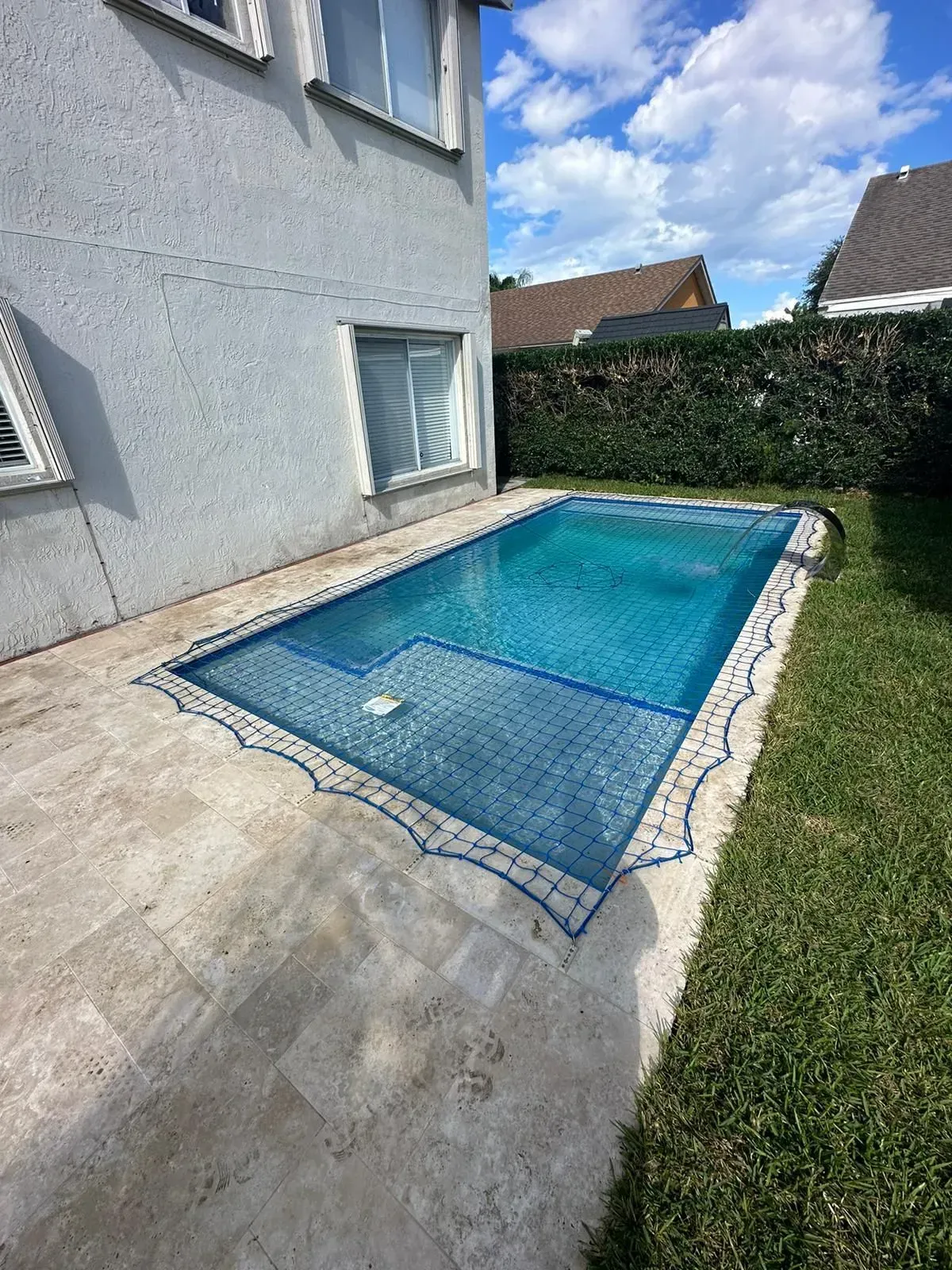 Small rectangular pool next to a light-colored house with blue water, surrounded by light stone and green grass.