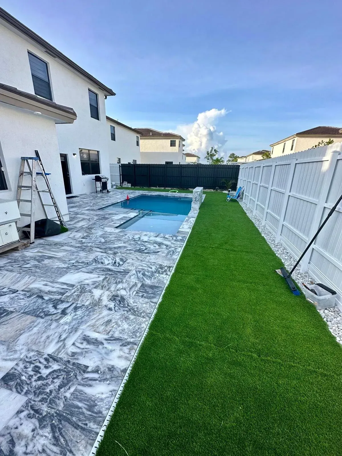 Backyard with a pool, patio, and green turf. White house and fence under a blue sky.