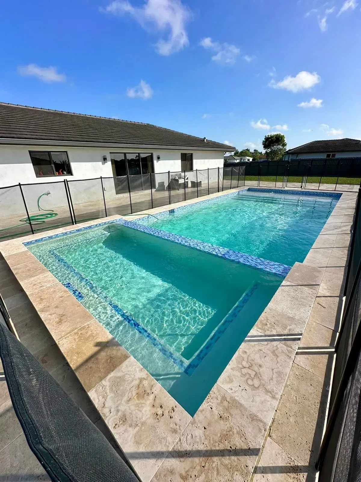 Rectangular swimming pool with light blue water and stone deck, against white house, under blue sky.