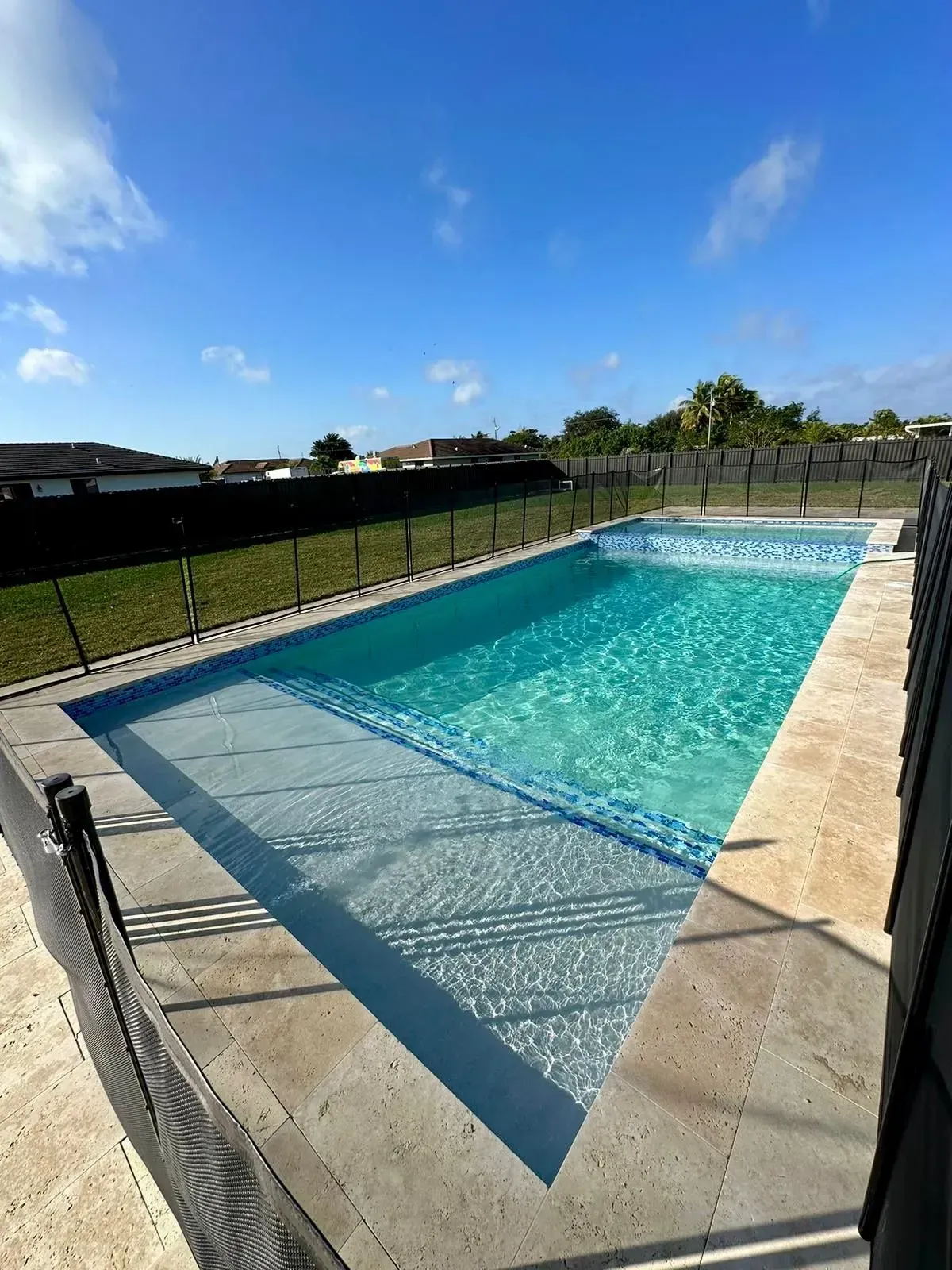 Rectangular swimming pool with clear water, blue tile trim, and gray concrete deck under a blue sky.