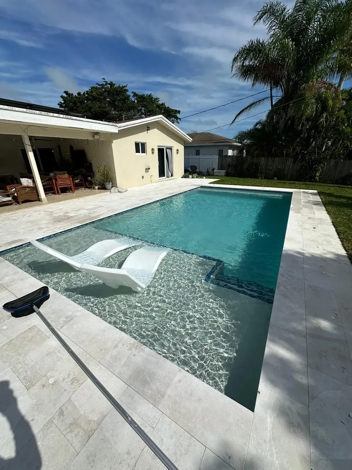 Swimming pool with white lounge chair, surrounded by a light-colored patio and a house under a blue sky.