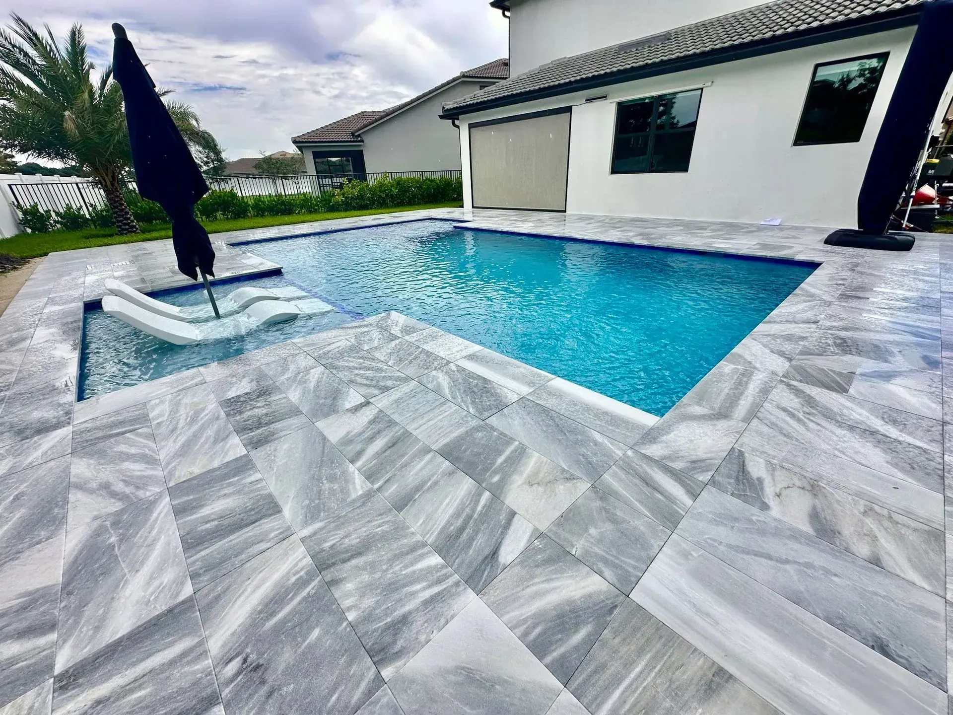 Swimming pool with gray stone tiles, blue water, and a white slide. A black umbrella is open beside it.