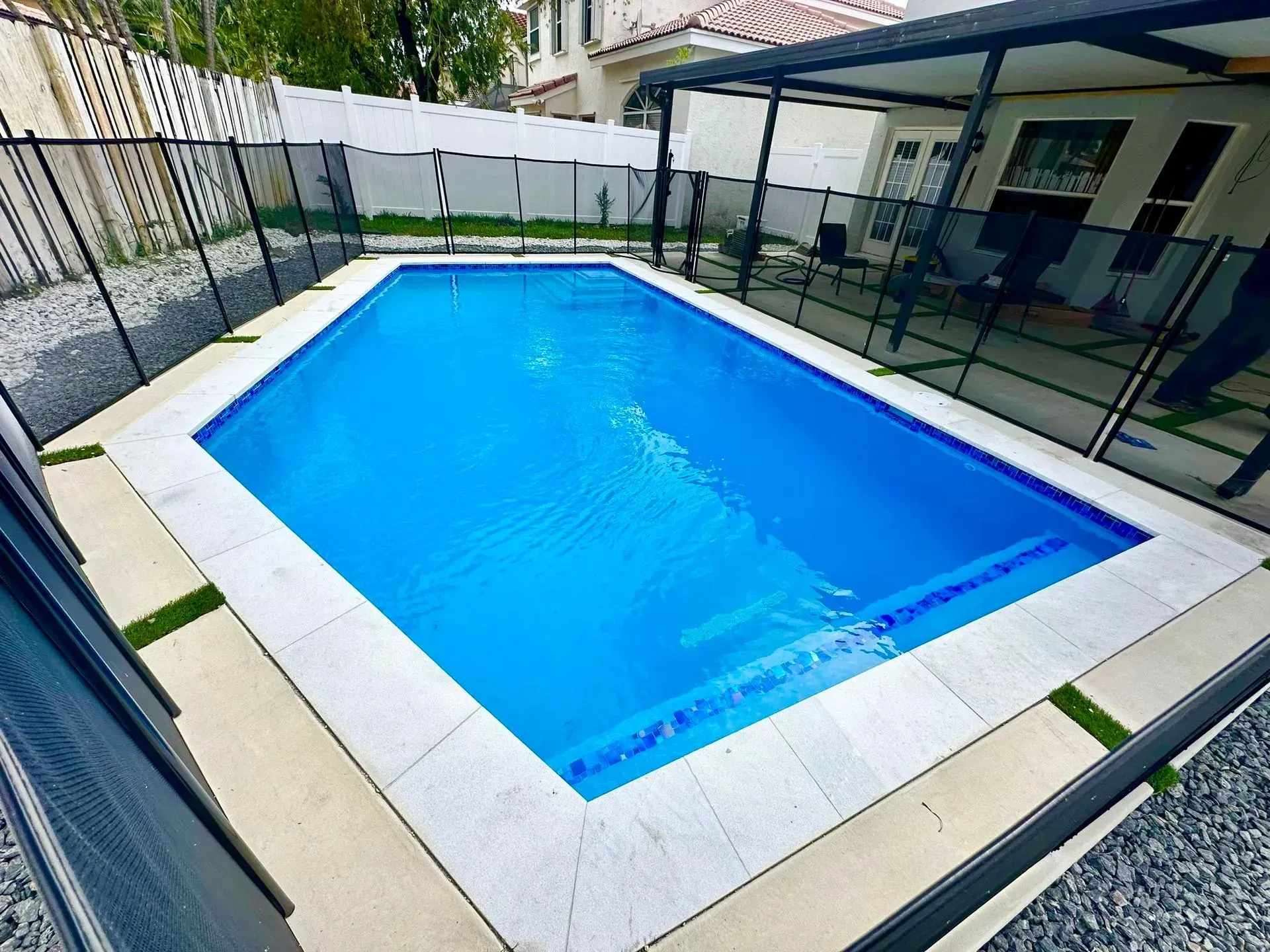 Rectangular blue pool surrounded by a white stone border, black safety fence, and a patio with a pergola.
