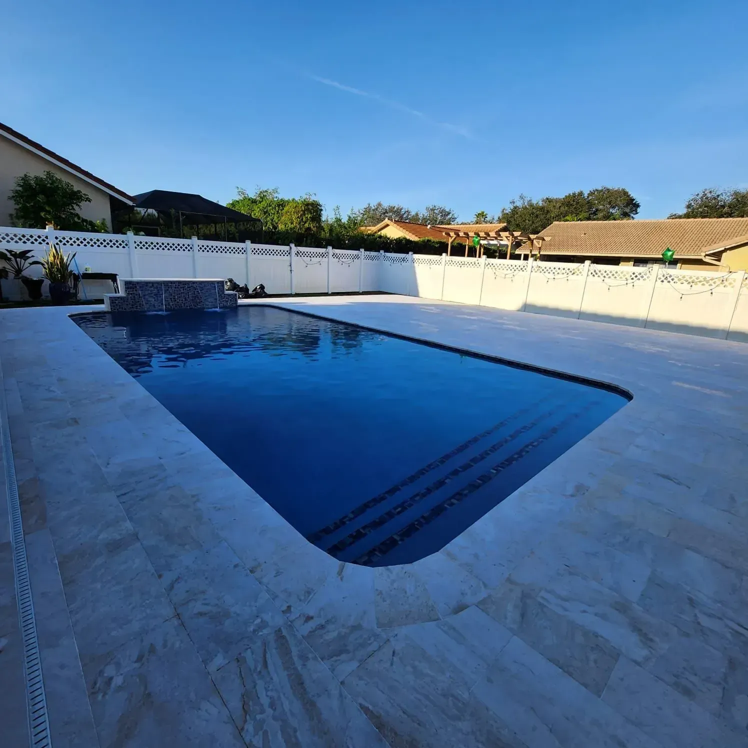 Rectangular pool surrounded by light-colored stone patio. Houses and a blue sky are in the background.