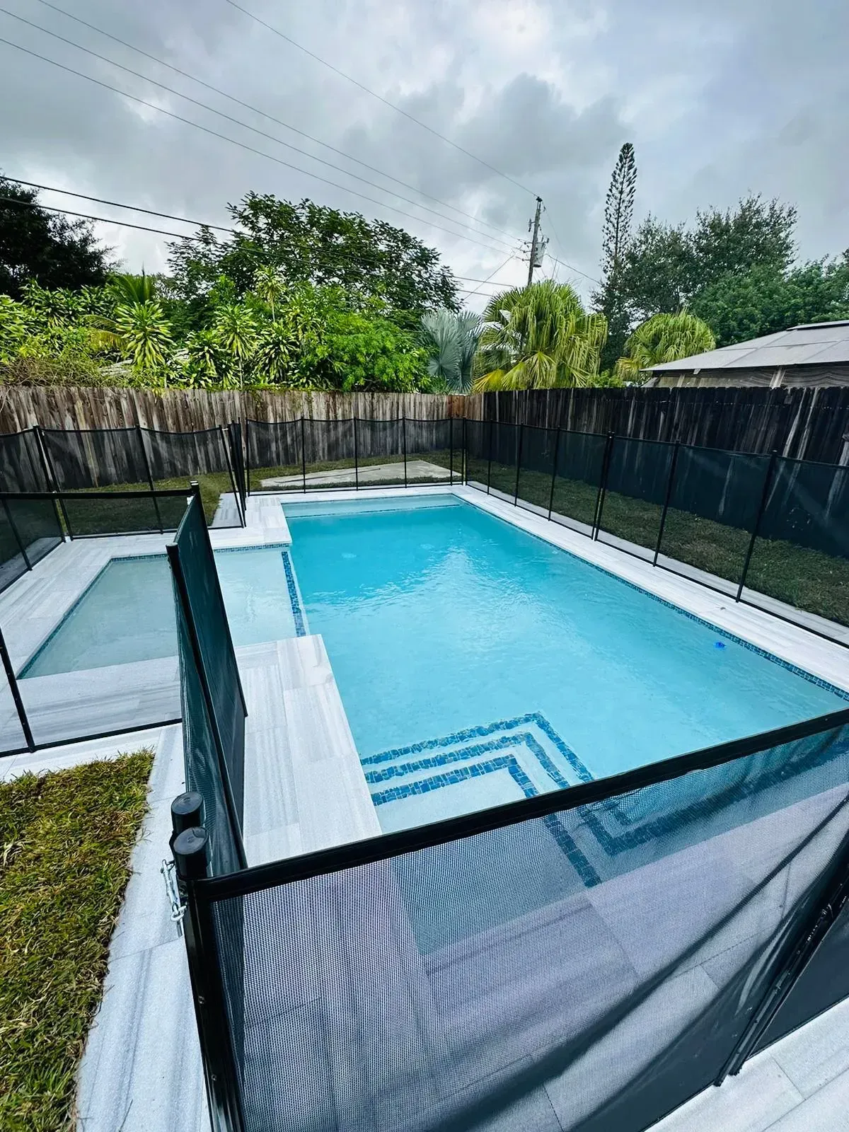 Swimming pool surrounded by a black mesh fence, white patio and green grass against a cloudy sky.