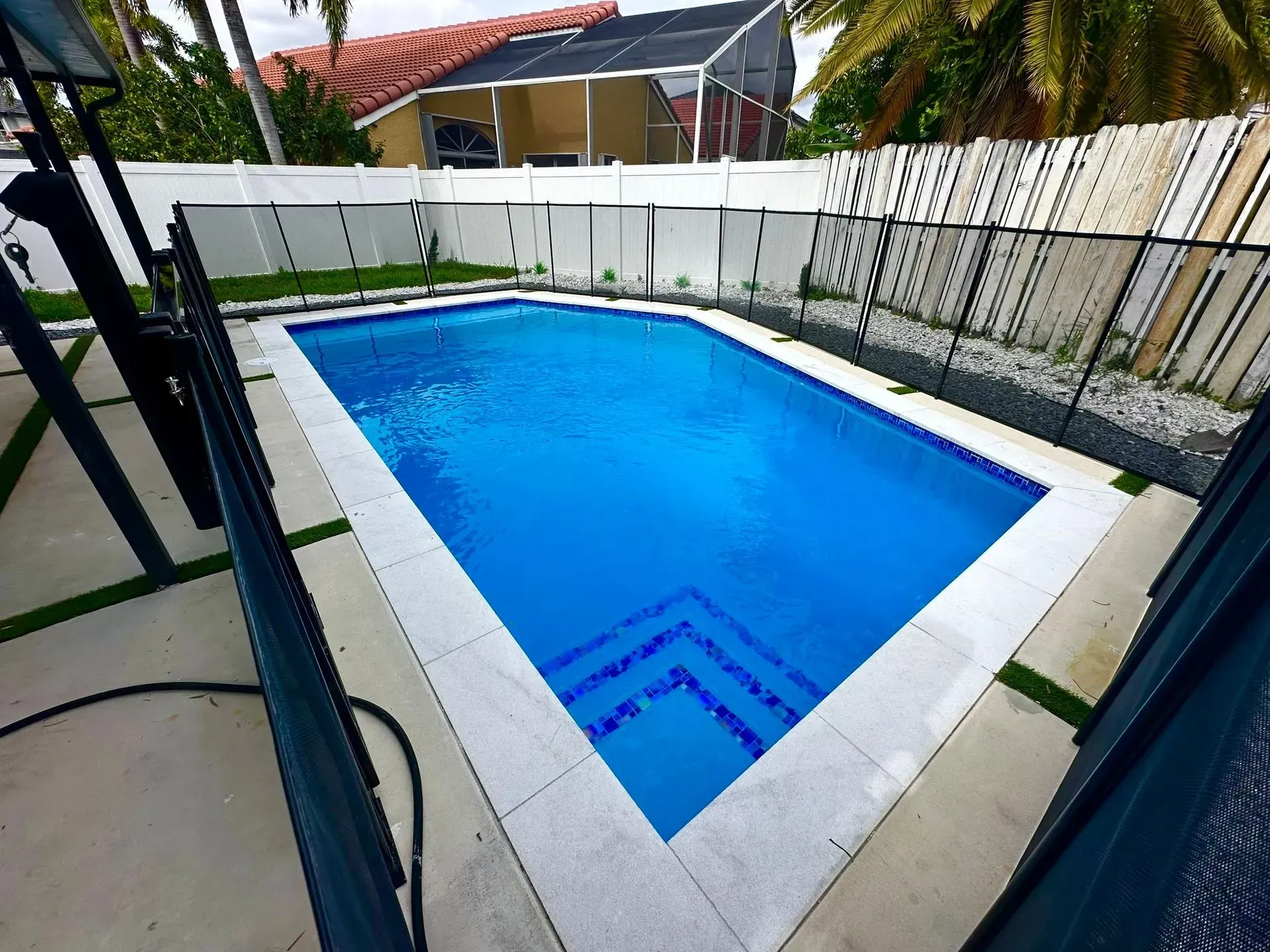 Rectangular swimming pool with blue water and steps, surrounded by a fence and a patio.