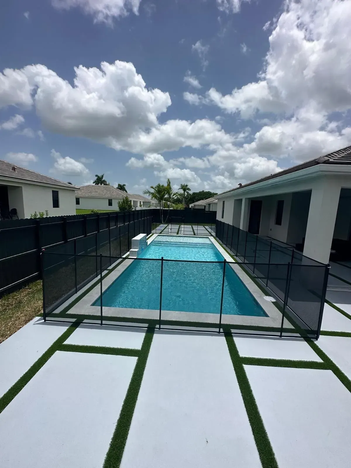 Rectangular pool with black safety fence, surrounded by white concrete and green artificial turf under a blue sky.