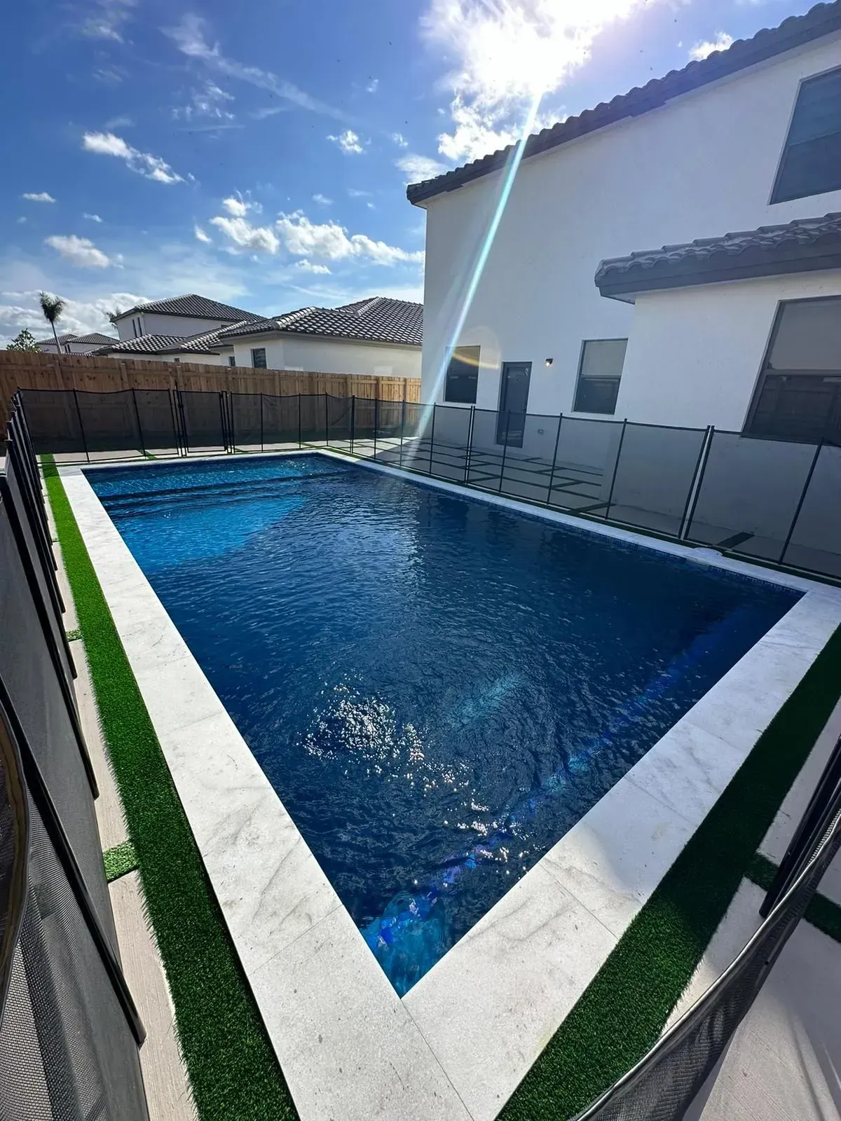 Rectangular pool with dark blue water, white border, and green turf, next to a white house.